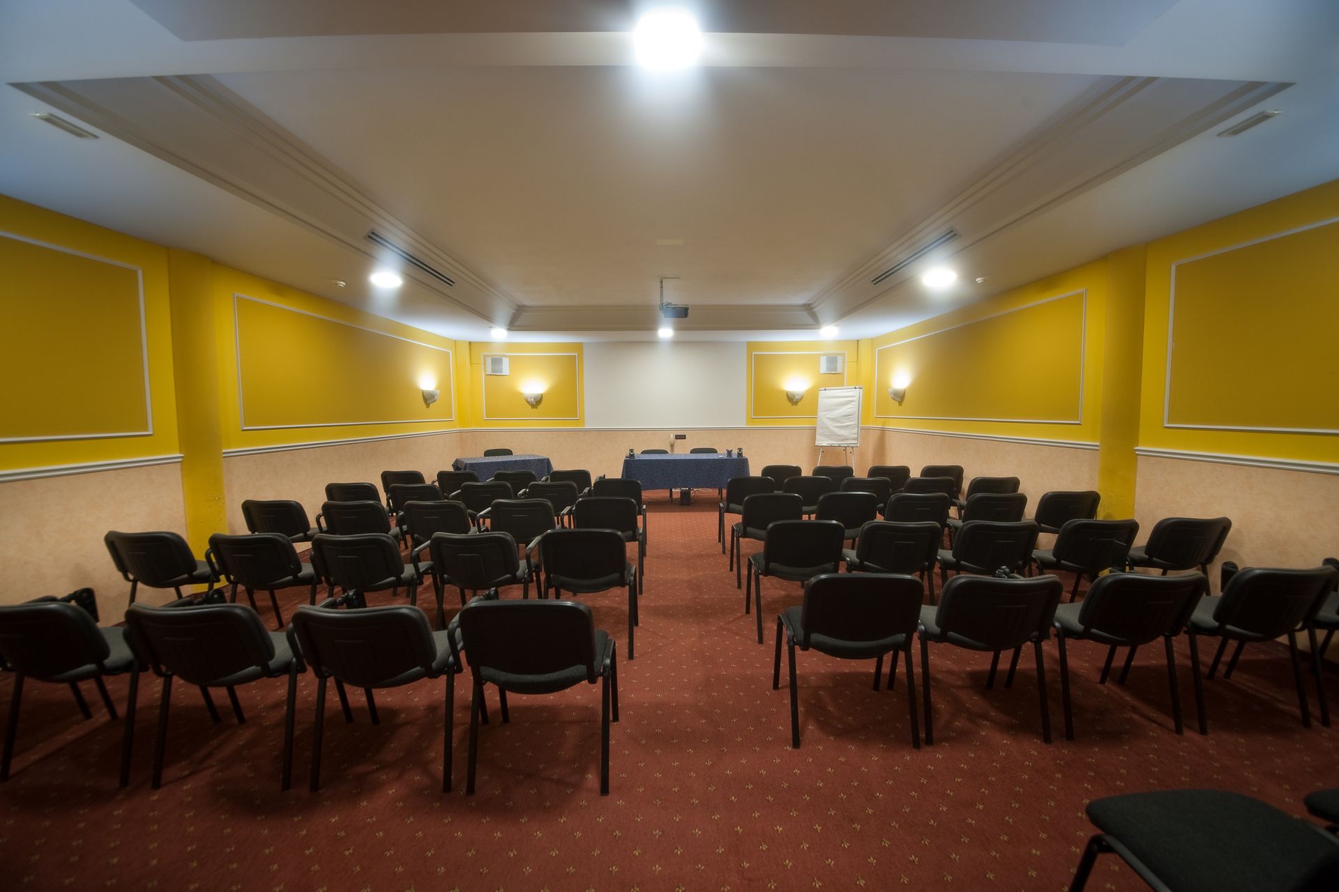 Konferenzraum mit Reihen schwarzer Stühle, die auf einen Tisch und eine Projektionsfläche ausgerichtet sind.