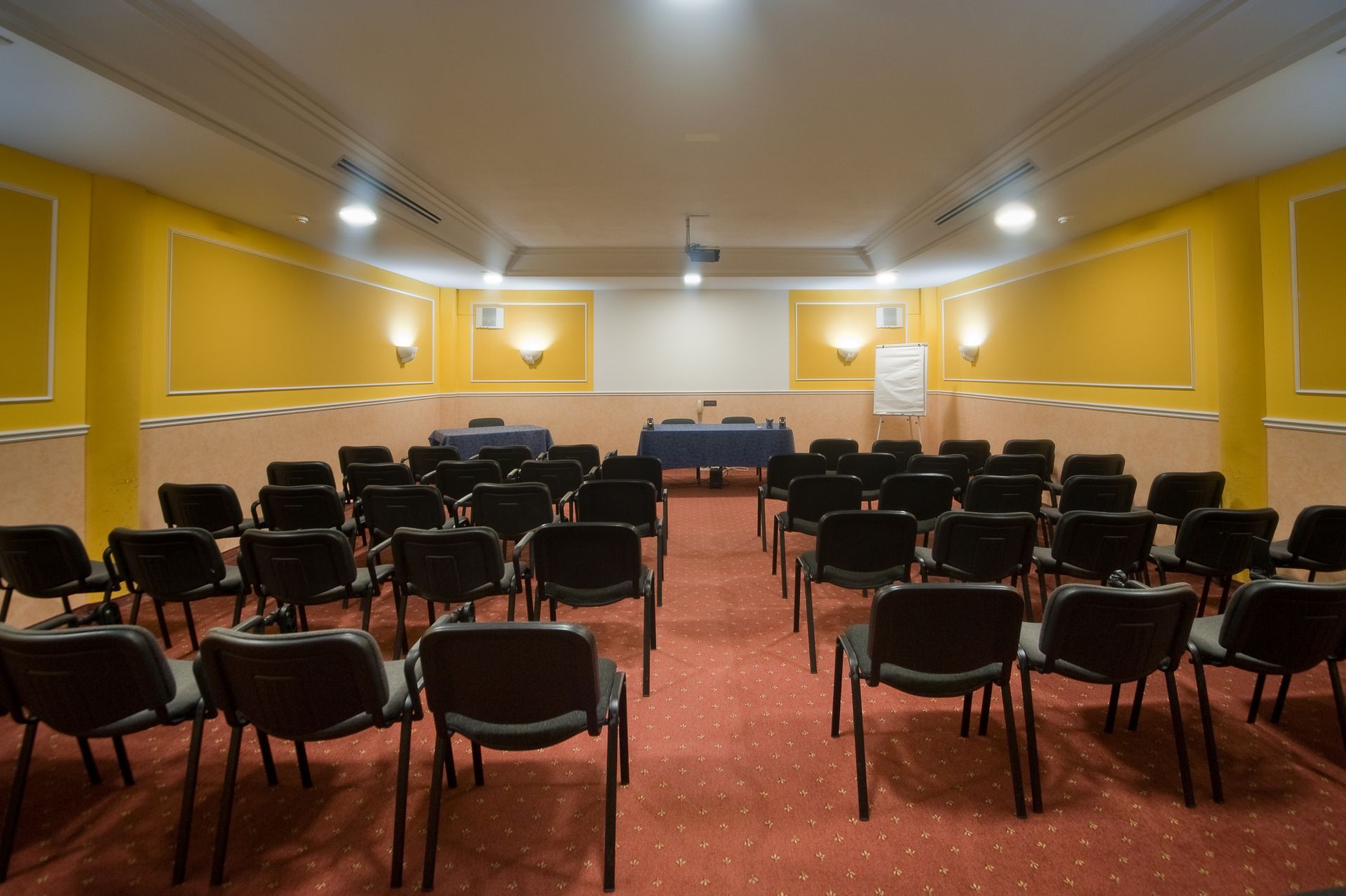 Sala riunioni con file di sedie nere di fronte a un tavolo e una lavagna bianca su un tappeto rosso.