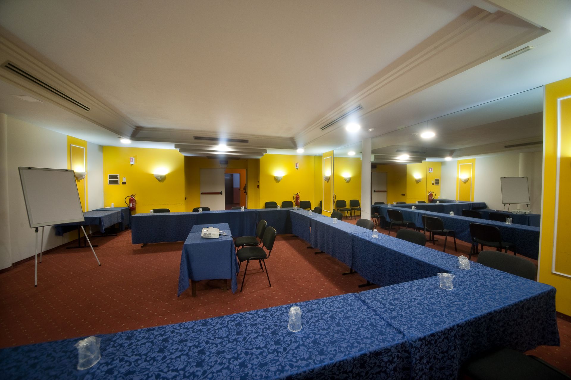 Sala conferenze allestita con tavoli a U, tovaglie blu e lavagne a fogli mobili. Pareti gialle, moquette rossa.
