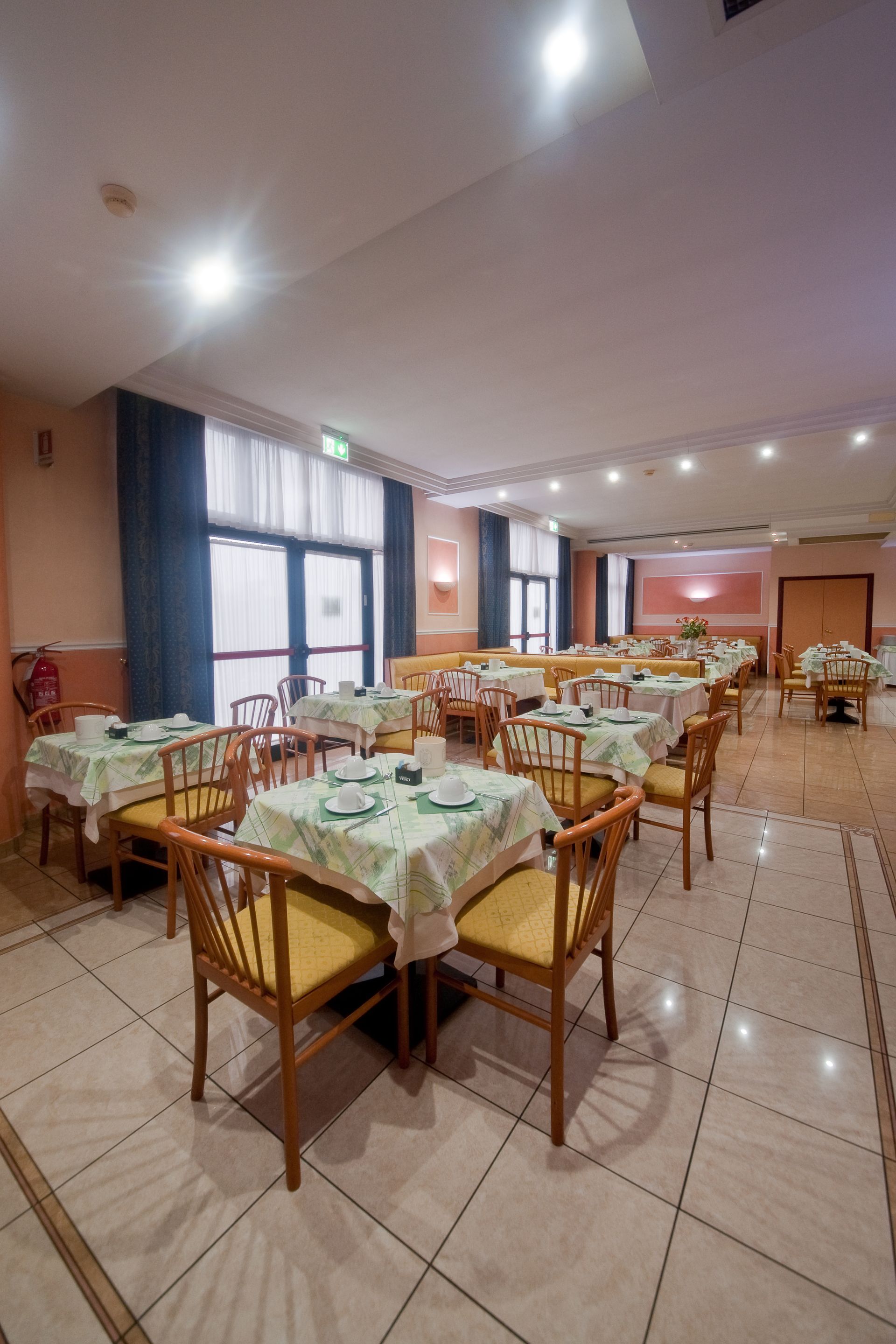 Sala da pranzo con tavoli apparecchiati per i pasti, sedie, pavimento piastrellato e finestre con tende scure.