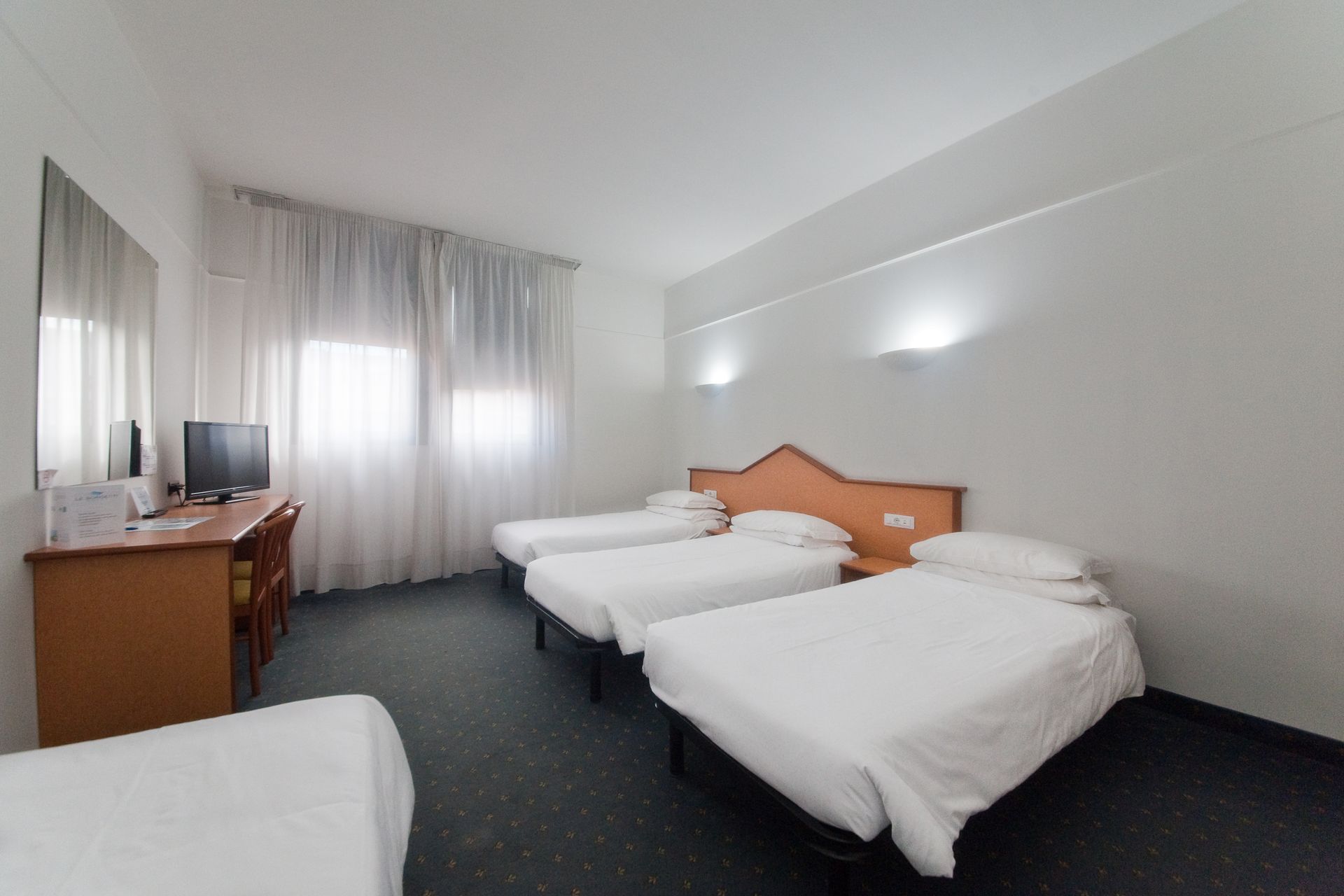 Camera d'albergo con quattro letti, scrivania, TV e finestra con tende trasparenti. Pareti bianche.