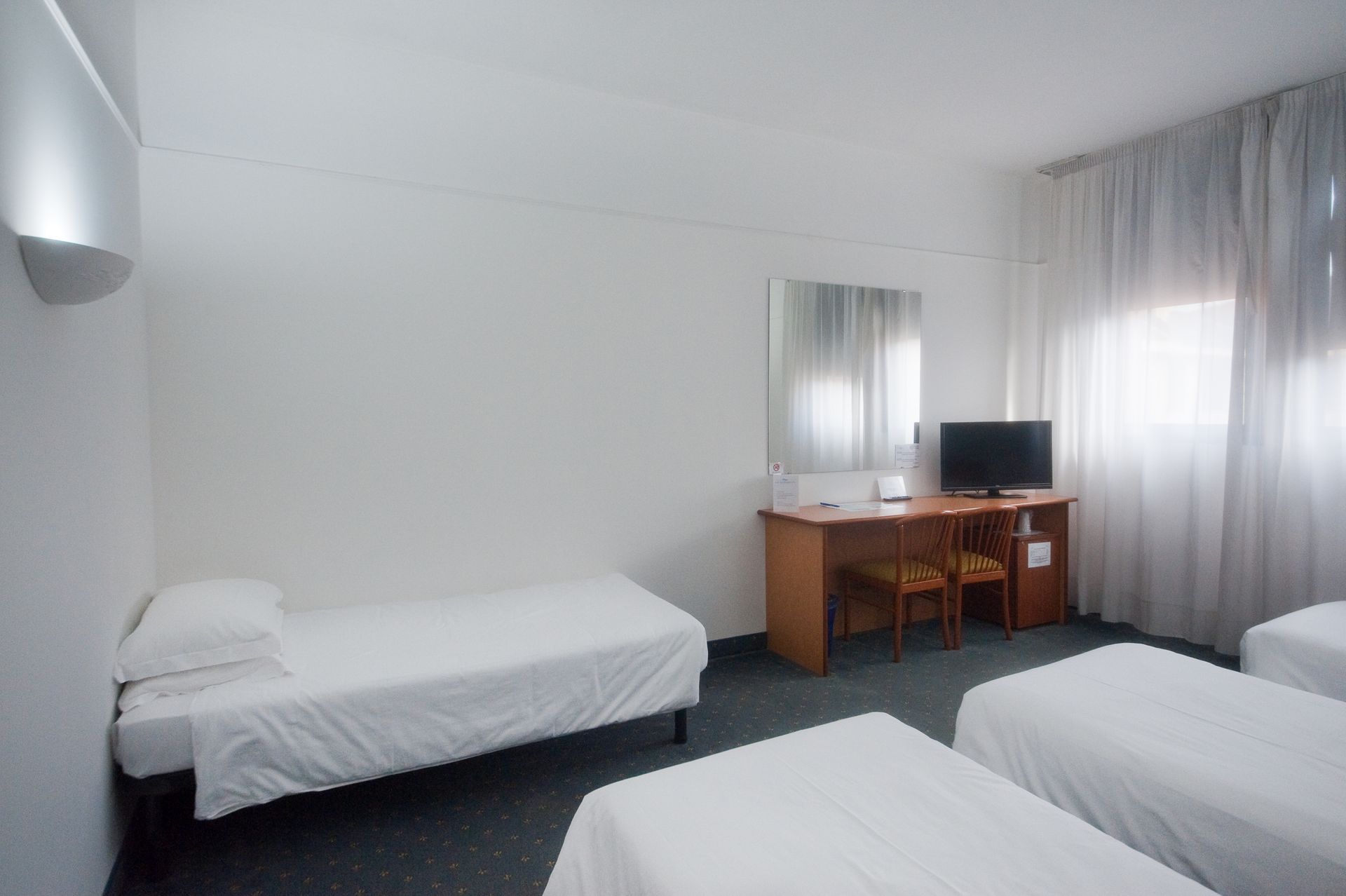 Camera d'albergo con quattro letti singoli, una scrivania con TV e una finestra con tende bianche.