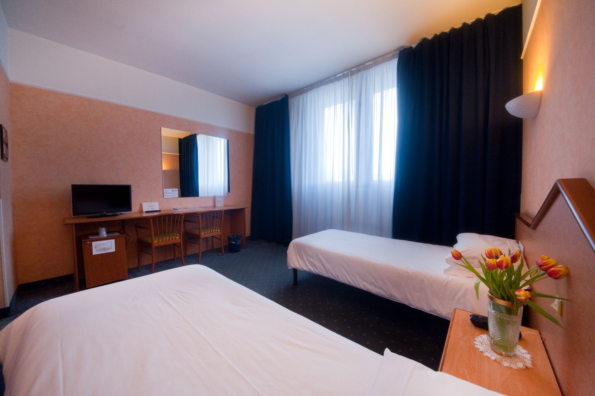 Hotelzimmer mit zwei Betten, kleinem Fernseher, Schreibtisch, Fenster mit transparenten und schwarzen Vorhängen, Blumenvase.