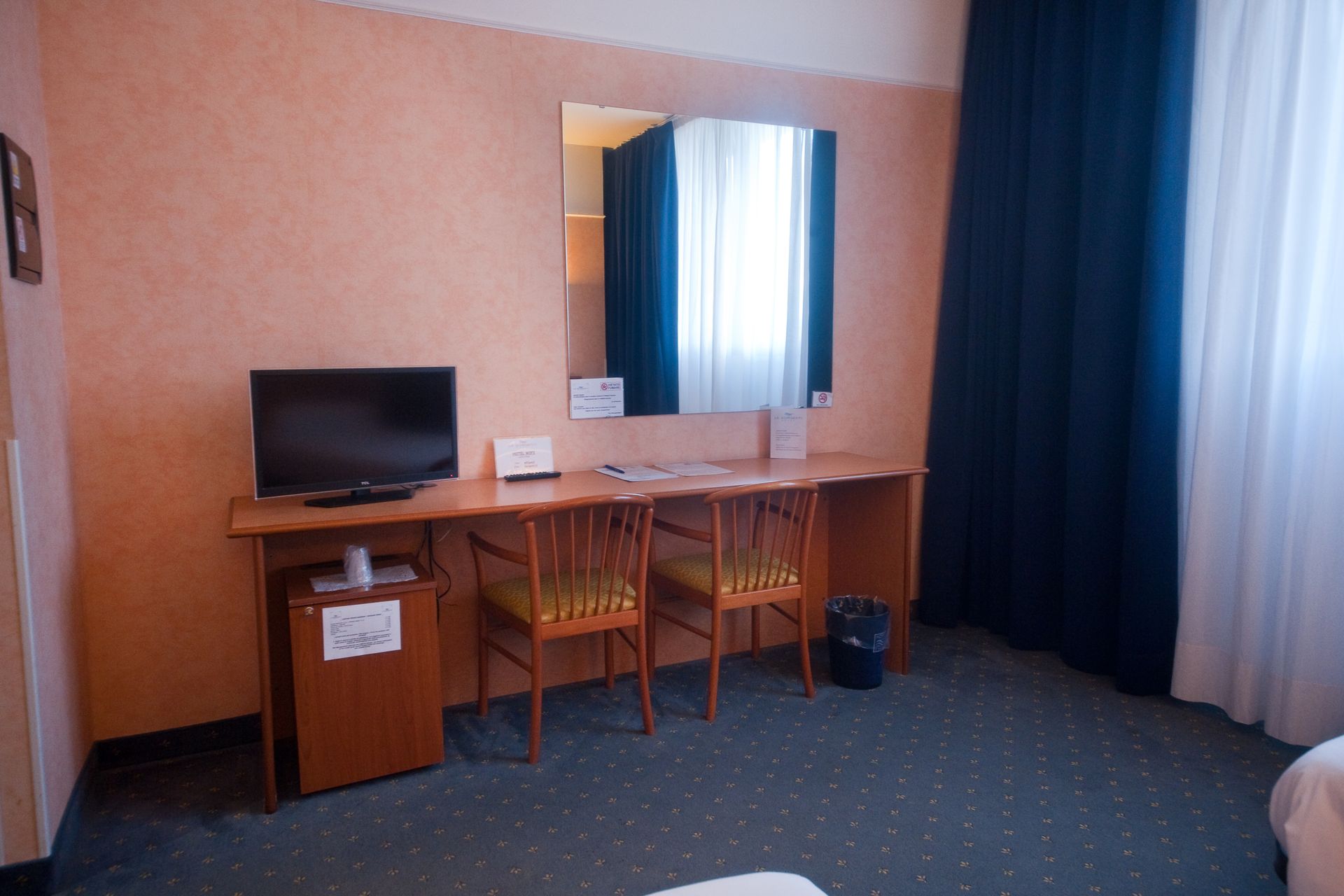 Interno di una camera d'albergo con scrivania, TV, sedie, specchio e finestra con tende.
