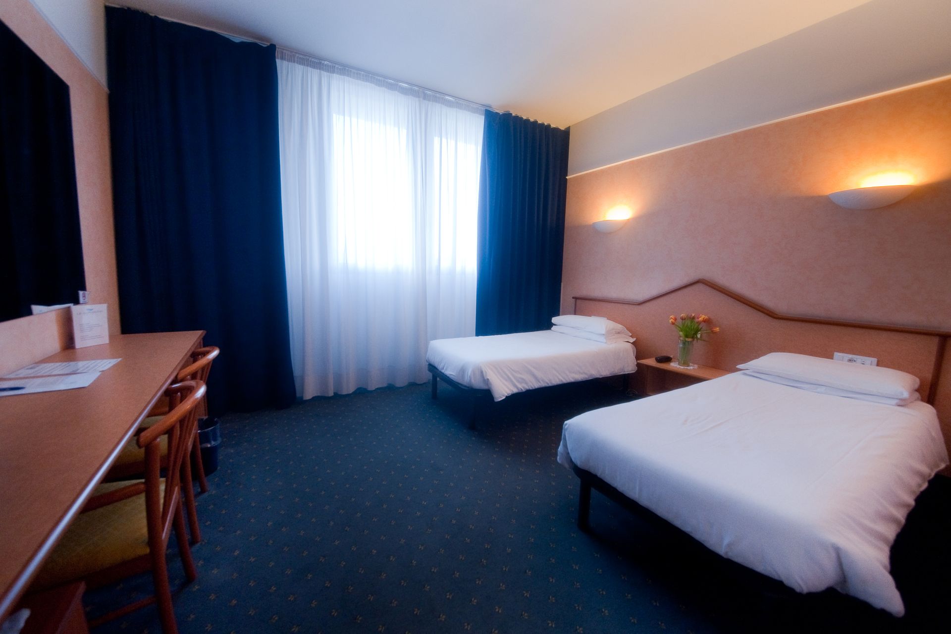 Camera d'albergo con due letti singoli, scrivania, finestra, tende blu scuro e pareti color pesca.