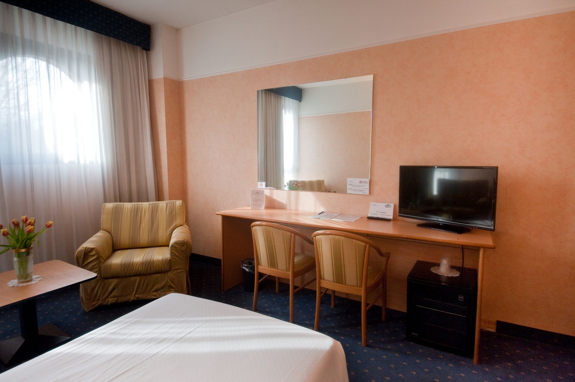 Camera d'albergo con letto, scrivania, sedie, poltrona, TV e finestra con tende; pareti color pesca e moquette blu.