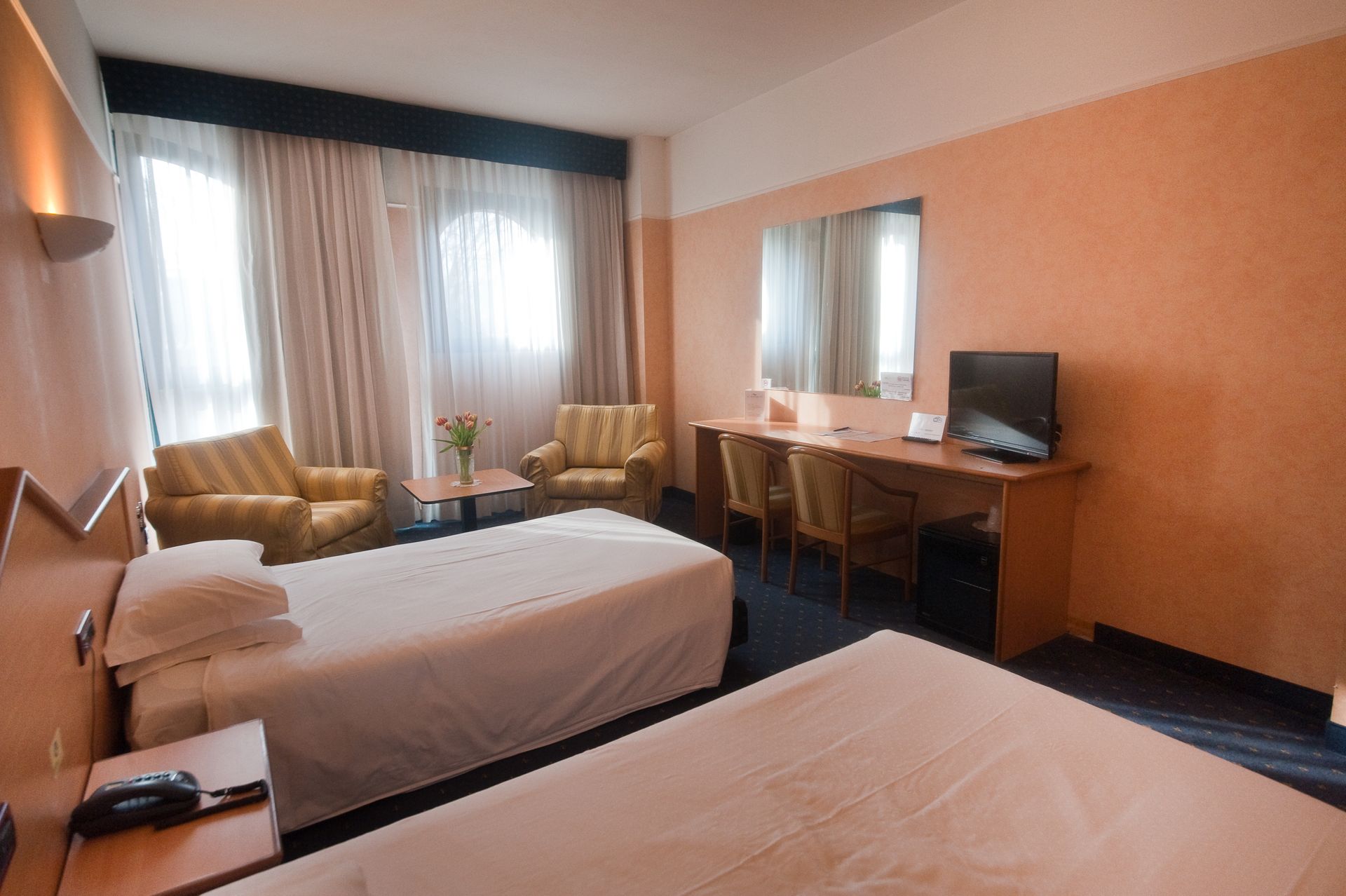 Camera d'albergo con due letti, sedie, scrivania, TV e finestra con tende.