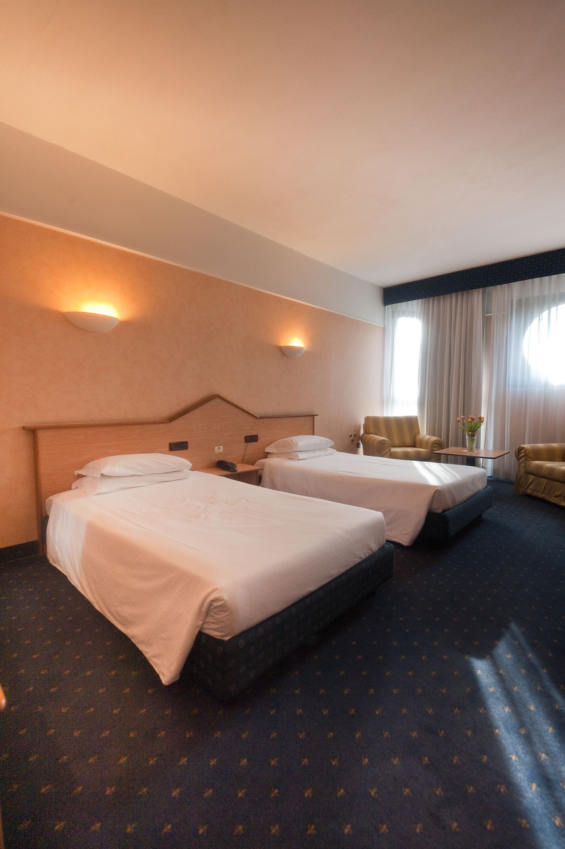 Camera d'albergo con due letti singoli, piccola area soggiorno e moquette blu.