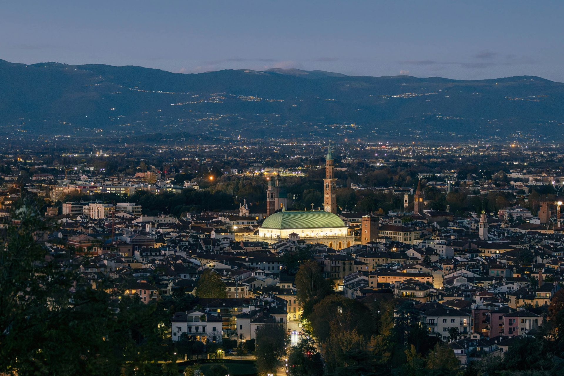 Stadtsilhouette in der Abenddämmerung, mit beleuchteten Gebäuden und Bergen im Hintergrund.