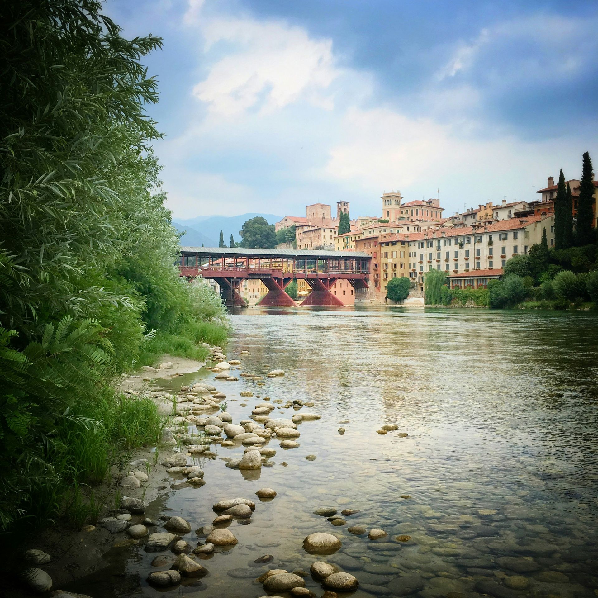 Ein Fluss mit einer Holzbrücke und farbenfrohen Gebäuden im Hintergrund unter einem bewölkten Himmel.