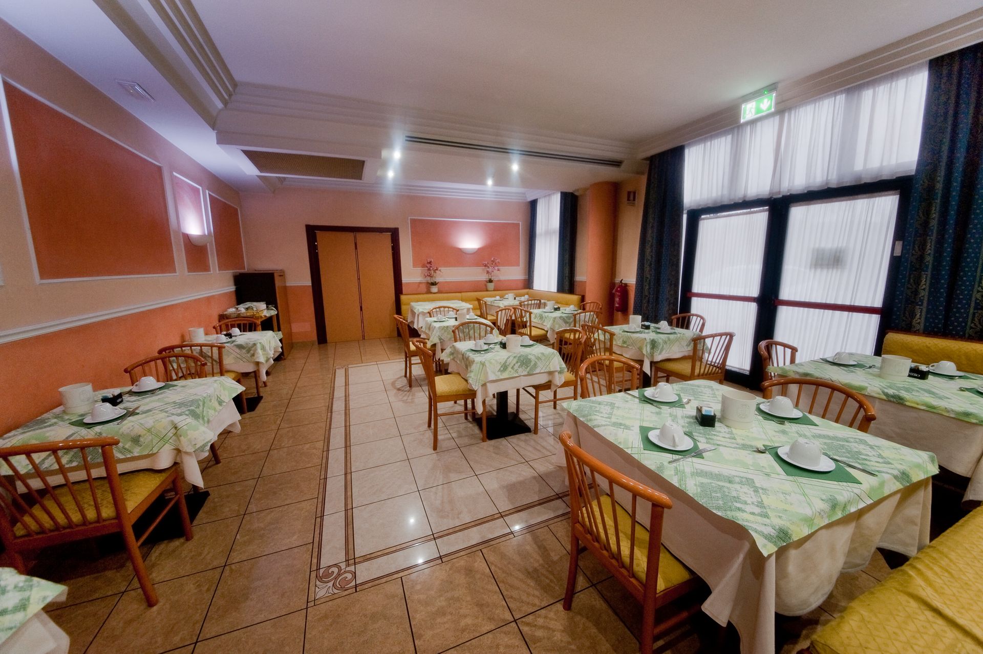 Sala da pranzo con tavoli apparecchiati per i pasti, pareti color arancione e crema, ampie finestre.