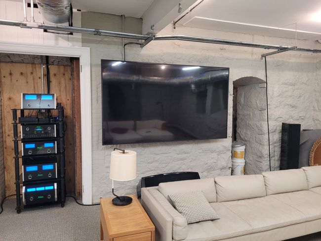 Loft living room with a large TV, beige sofa, lamp, and equipment rack against exposed brick and concrete.