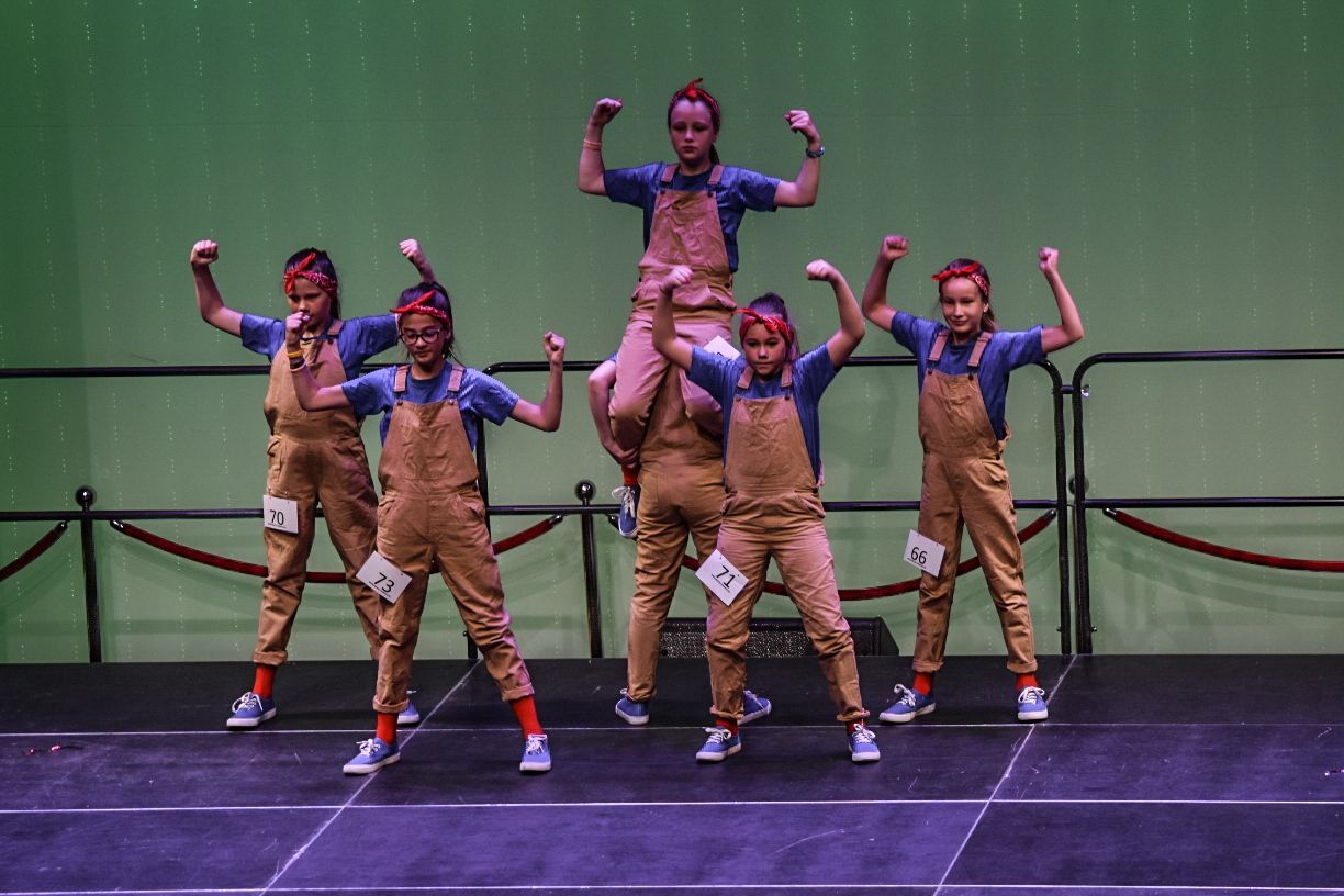 Een groep jonge meisjes in overalls danst op een podium.