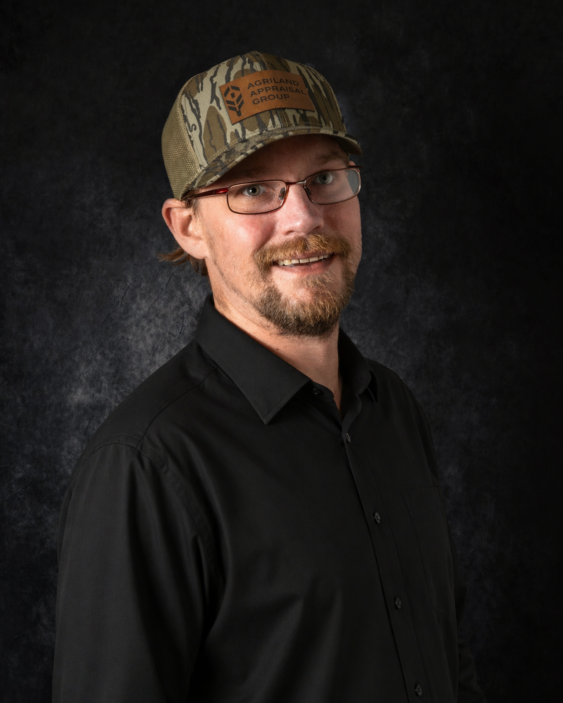 Man wearing glasses and camouflage hat smiles, gray collared shirt, dark background.