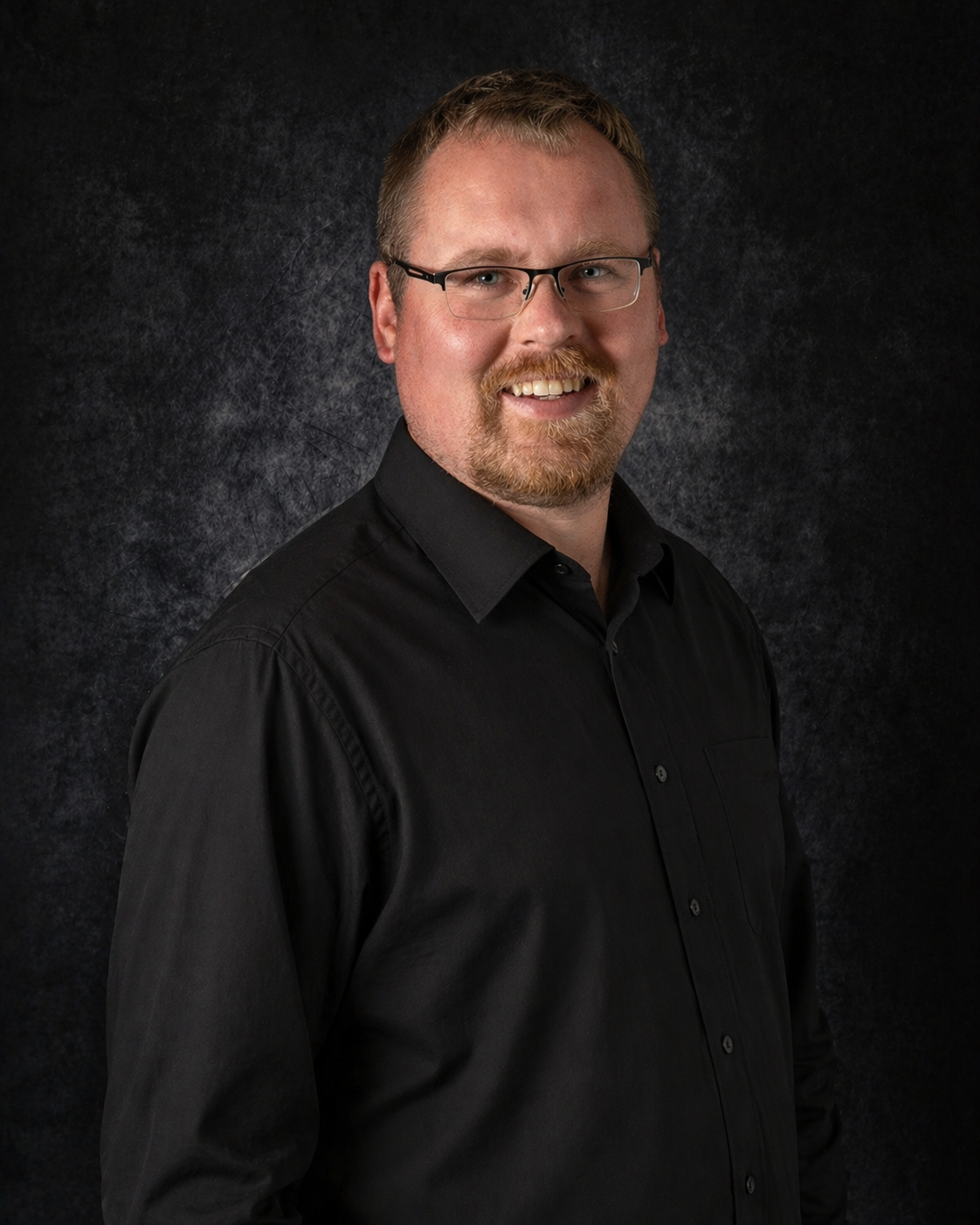 Man with glasses and beard wearing a vest over patterned shirt, smiling against a dark backdrop.