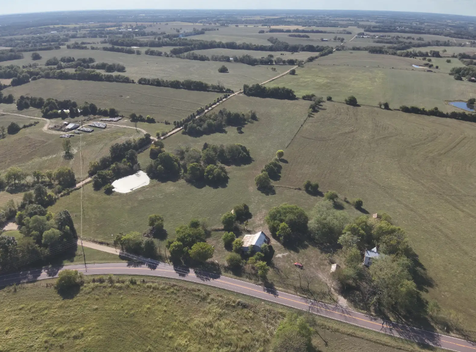 Aerial view of a rural landscape with a small house, a pond, trees, open fields, and a paved road.