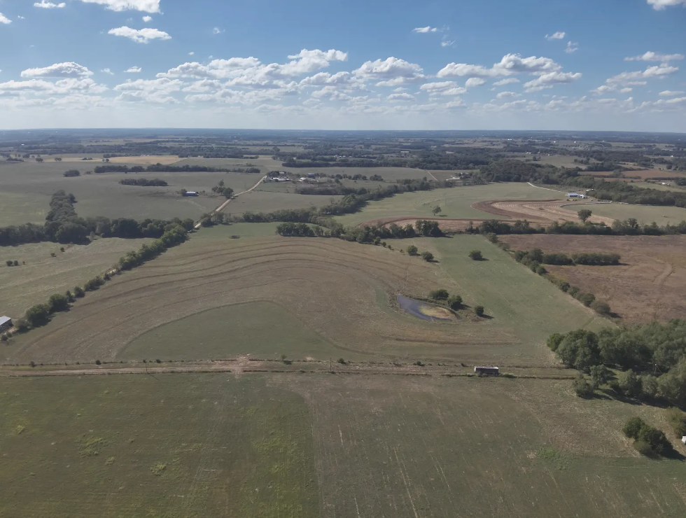 An aerial view of a sprawling landscape featuring grassy fields, curved agricultural rows, and a small pond under a blue sky.
