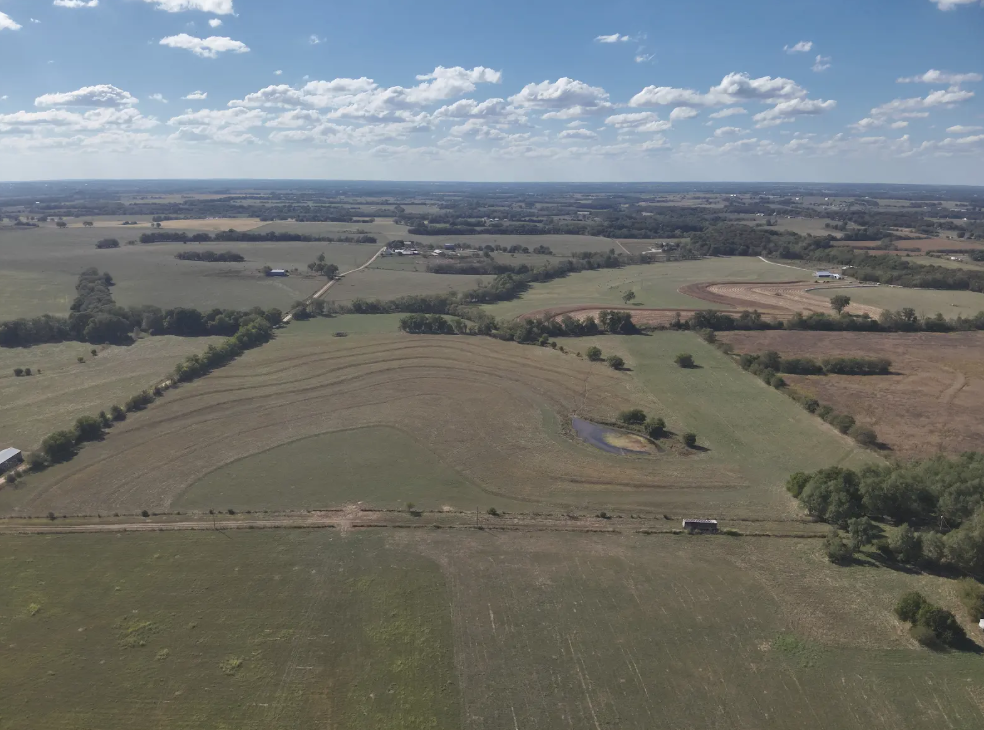 An aerial view of a large, rural landscape featuring open fields, scattered trees, and a small pond under a cloudy sky.