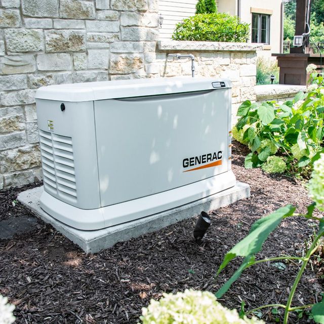A Generac home generator on a concrete pad next to a brick wall and landscaping.