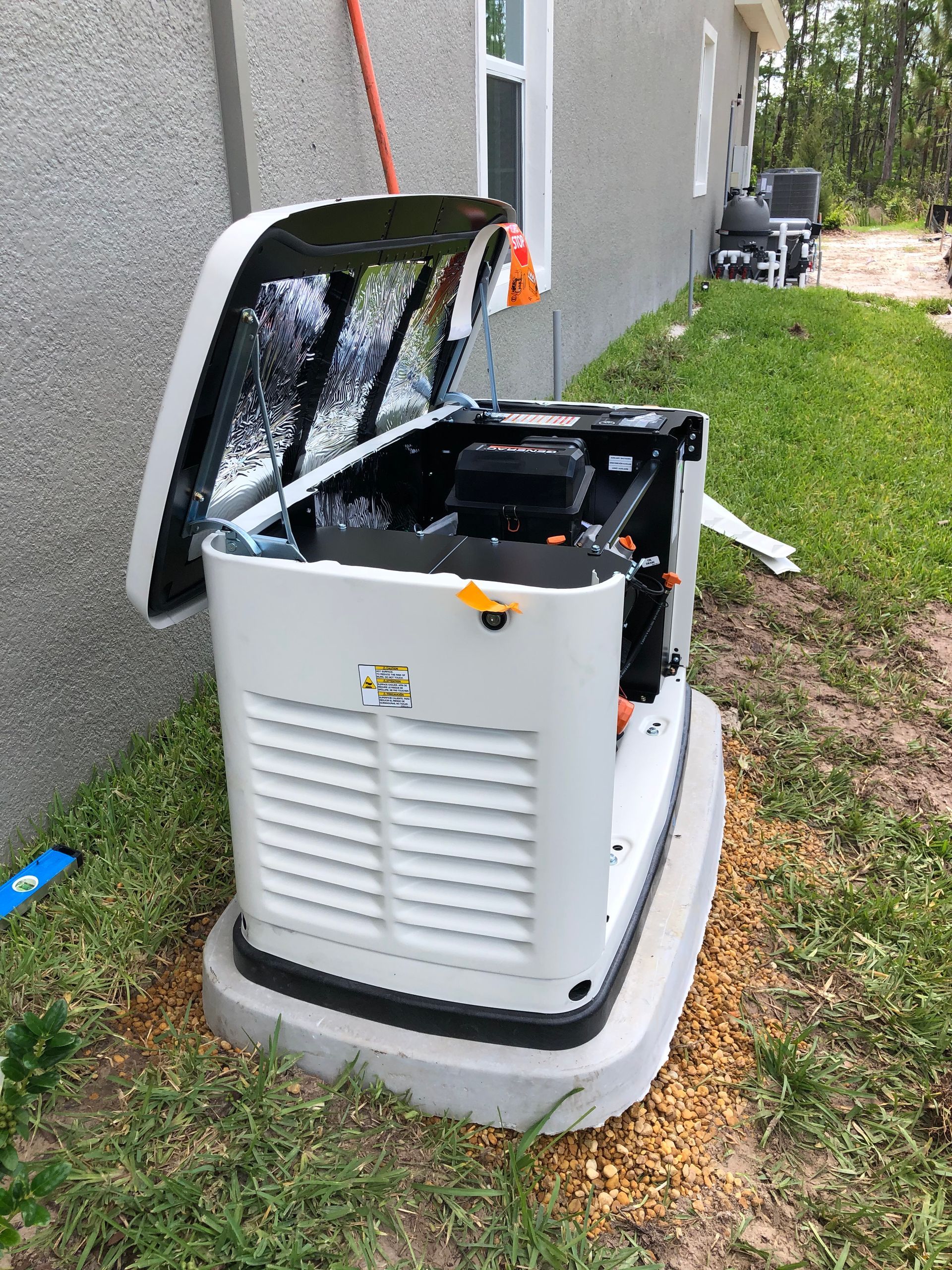 Generator with open top, white and black, on a concrete pad next to a house with a window, grass and trees.