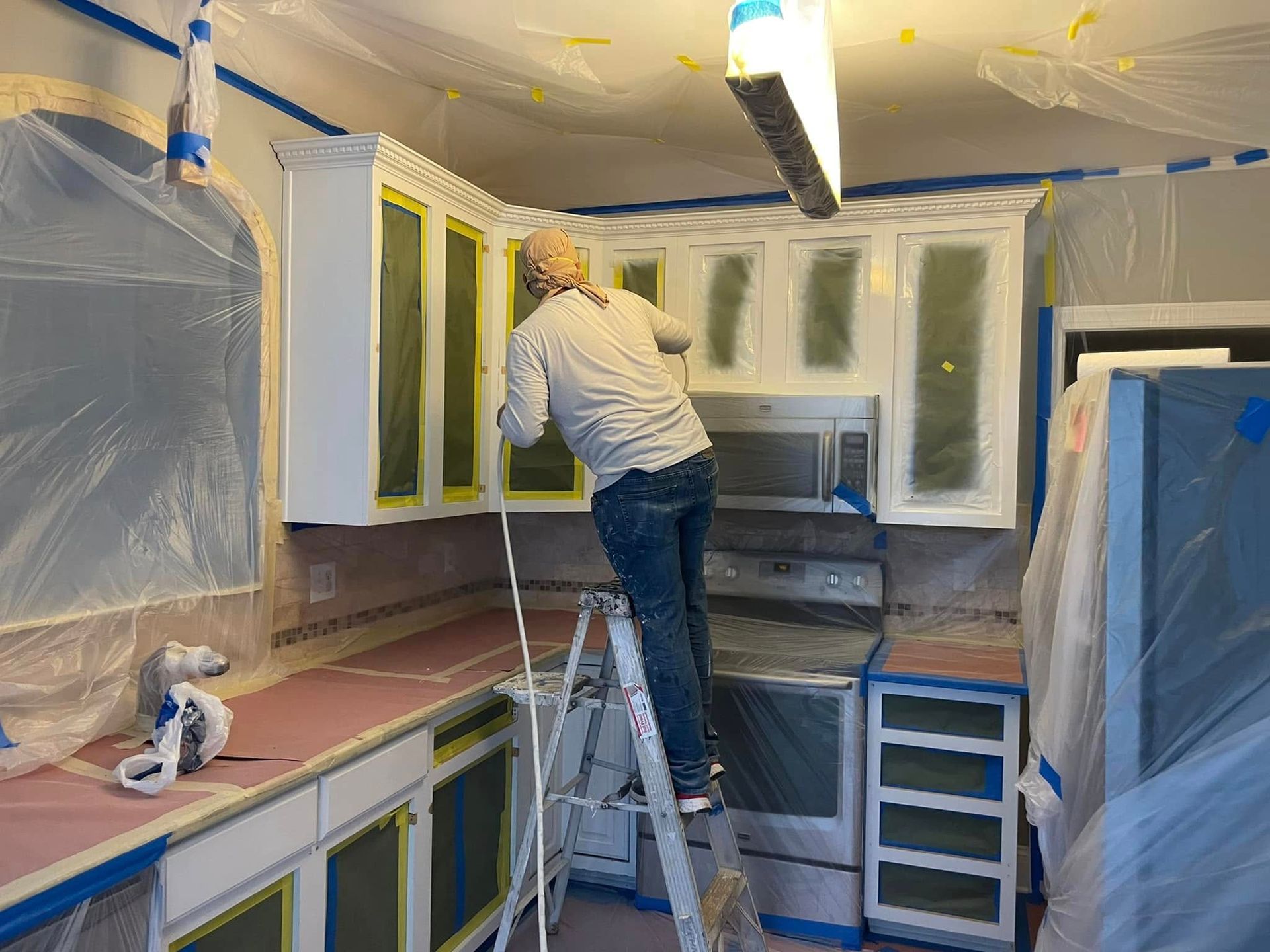 Person painting kitchen cabinets while standing on a step ladder; room covered with masking plastic.