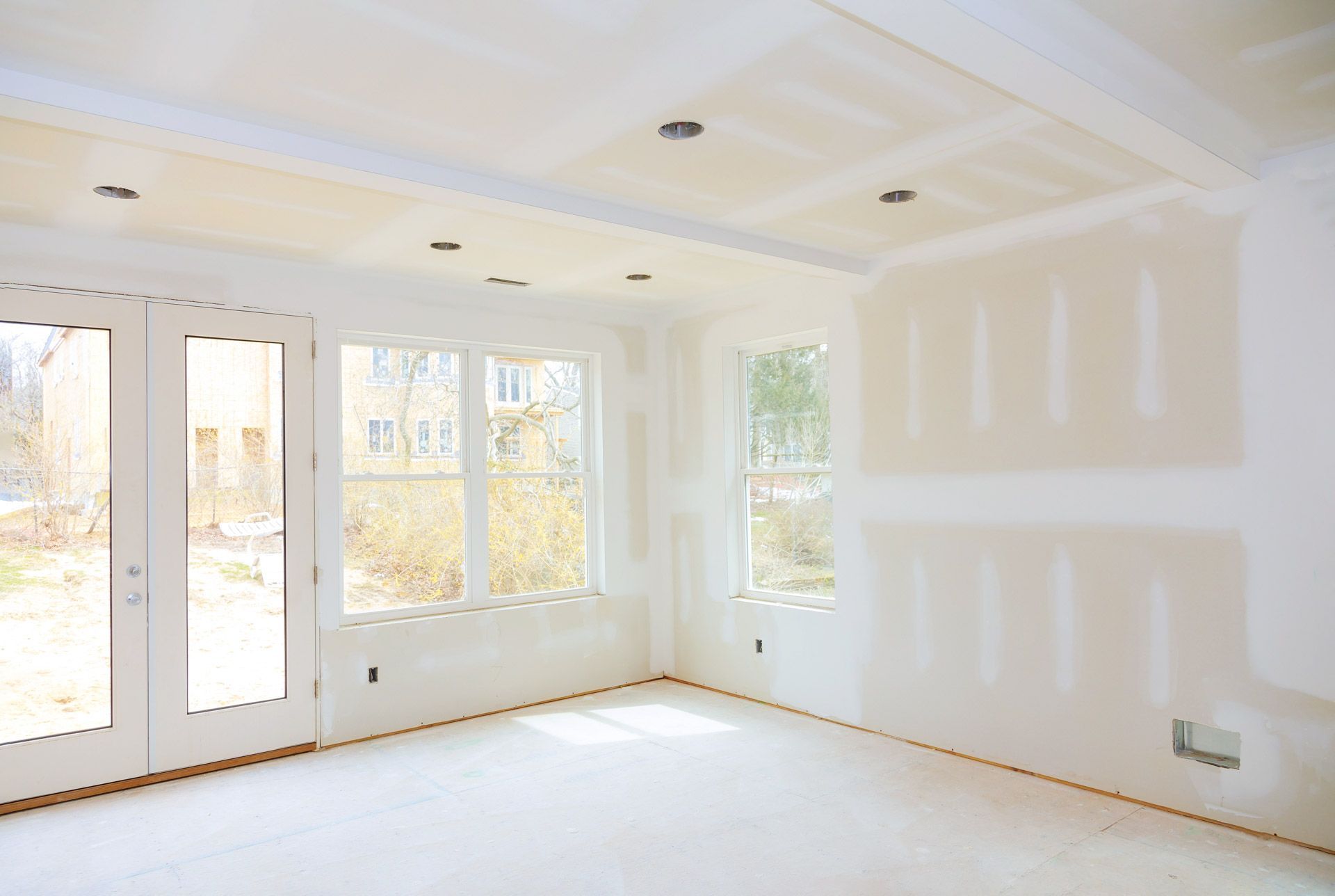 Bright empty living room with white walls, large windows, and unfinished drywall patches.