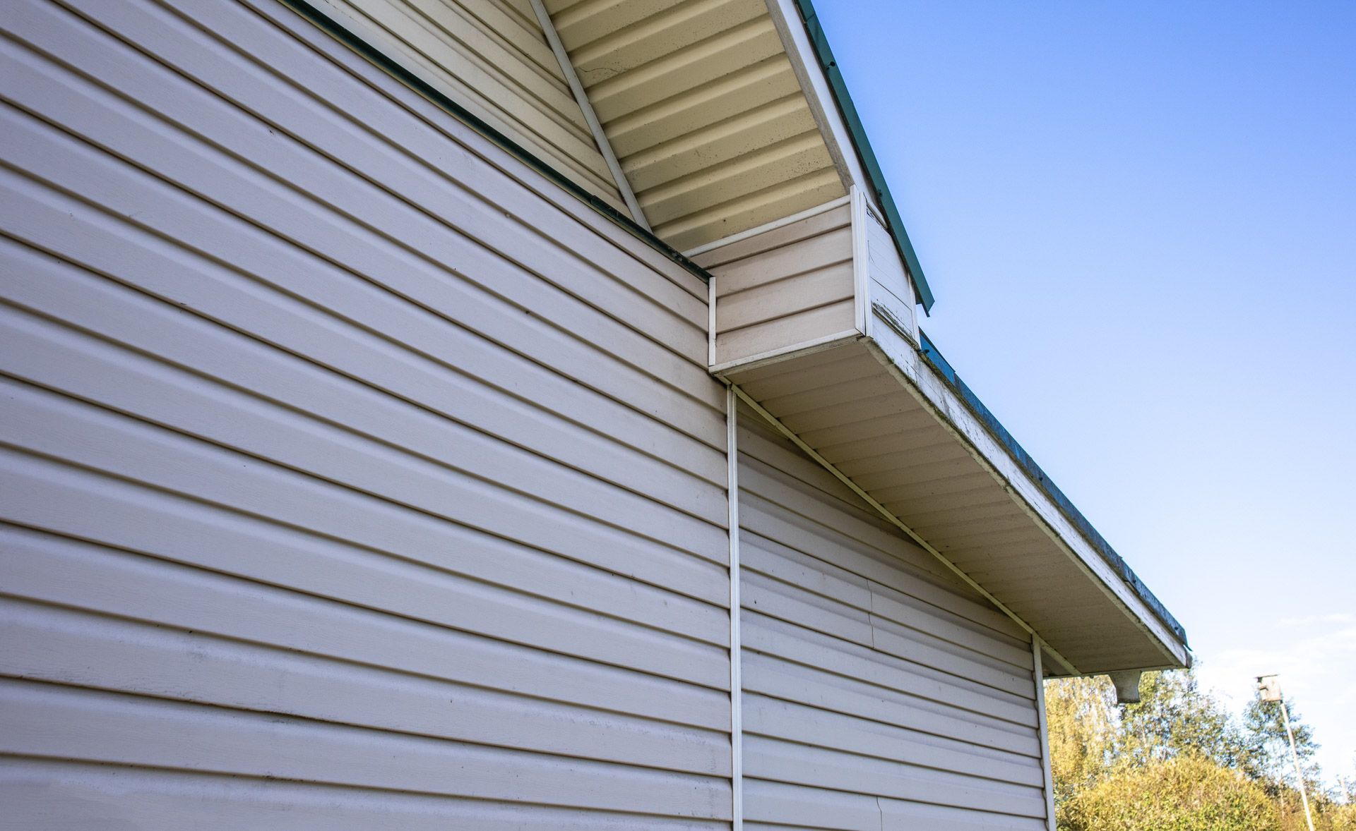 Close-up of light gray vinyl house siding and soffit under a roof eave against a blue sky.