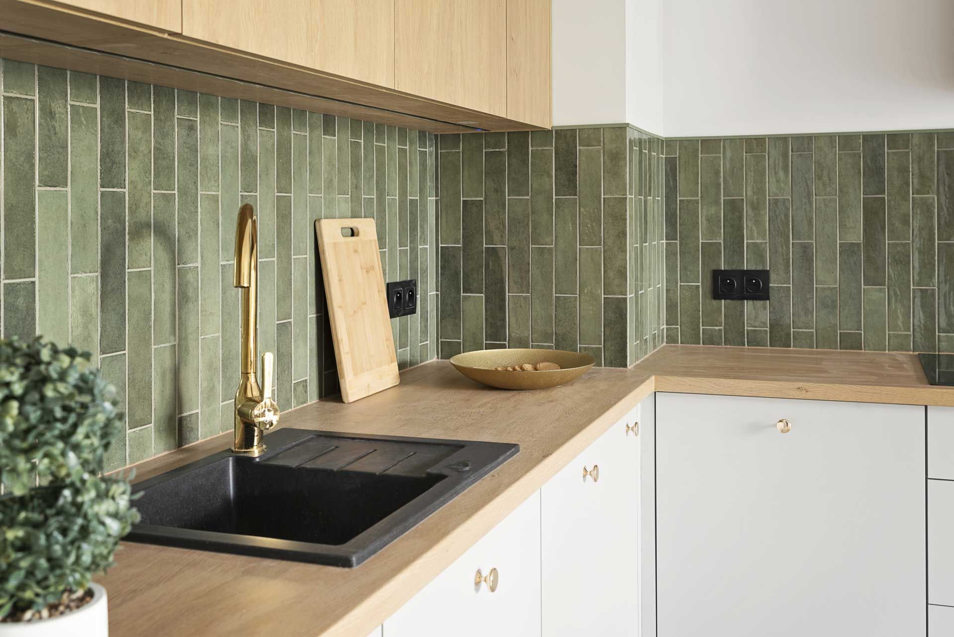 Modern kitchen corner with black sink, brass faucet, green tile backsplash, and light wood countertops