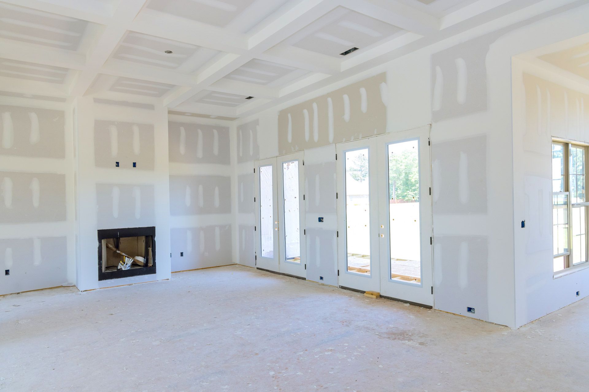 Bright unfinished living room with white drywall, fireplace, and large windows overlooking a yard