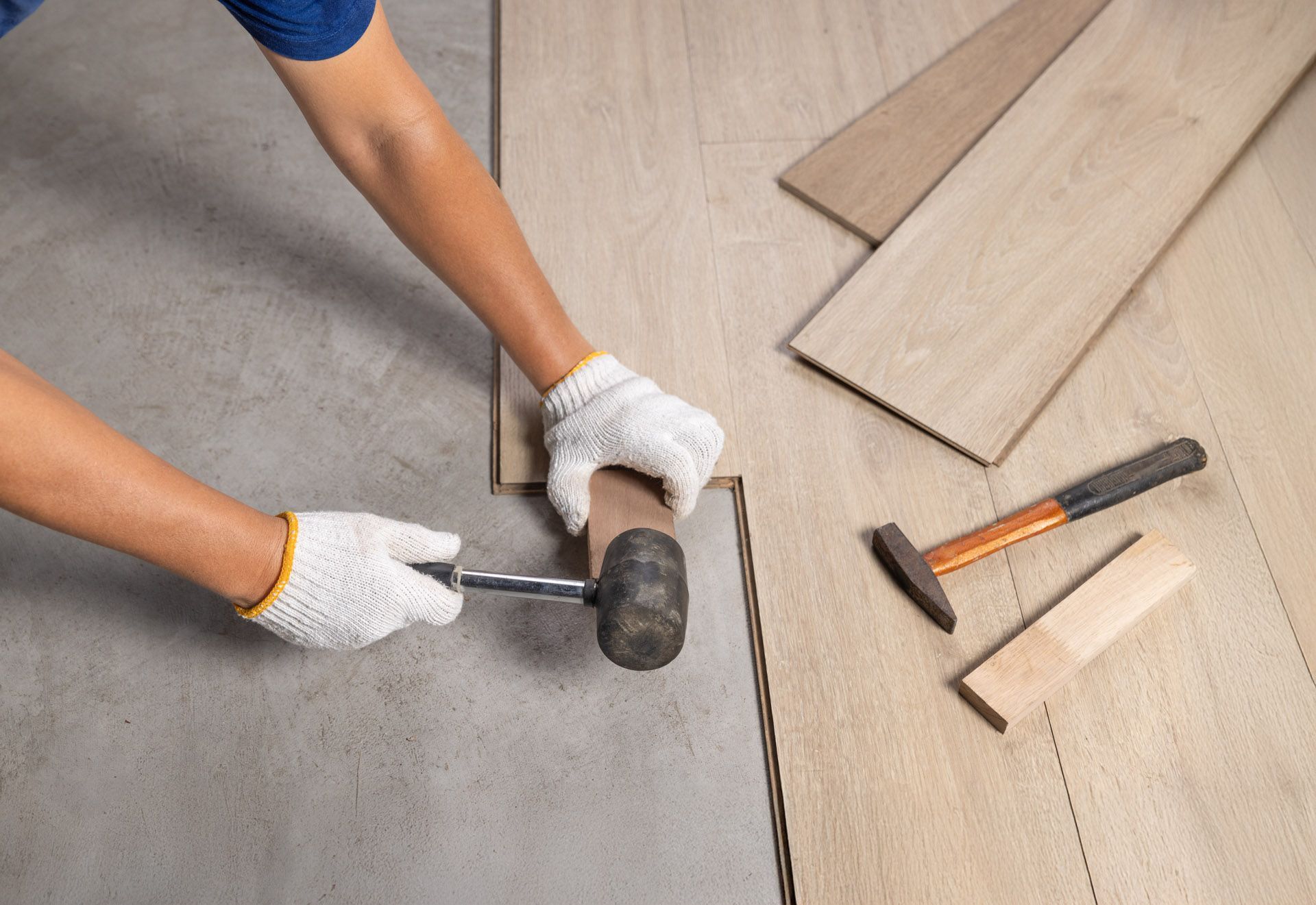 Gloved hands installing wood flooring with a mallet and tapping block beside unfinished planks