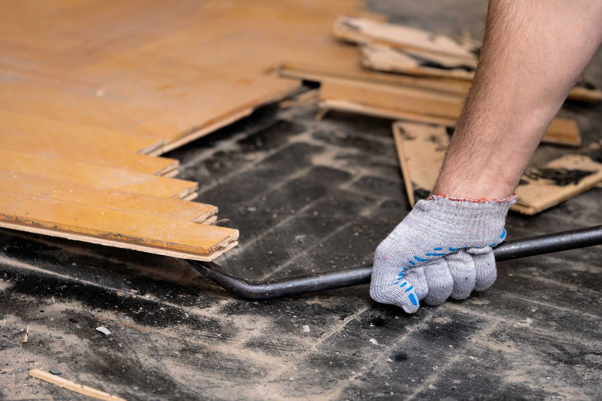 Person prying up old wooden flooring with a crowbar, revealing black underlayment.