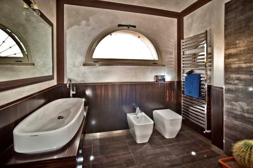 Bagno moderno con vasca freestanding, due WC bianchi, pareti in legno scuro e finestre ad arco.