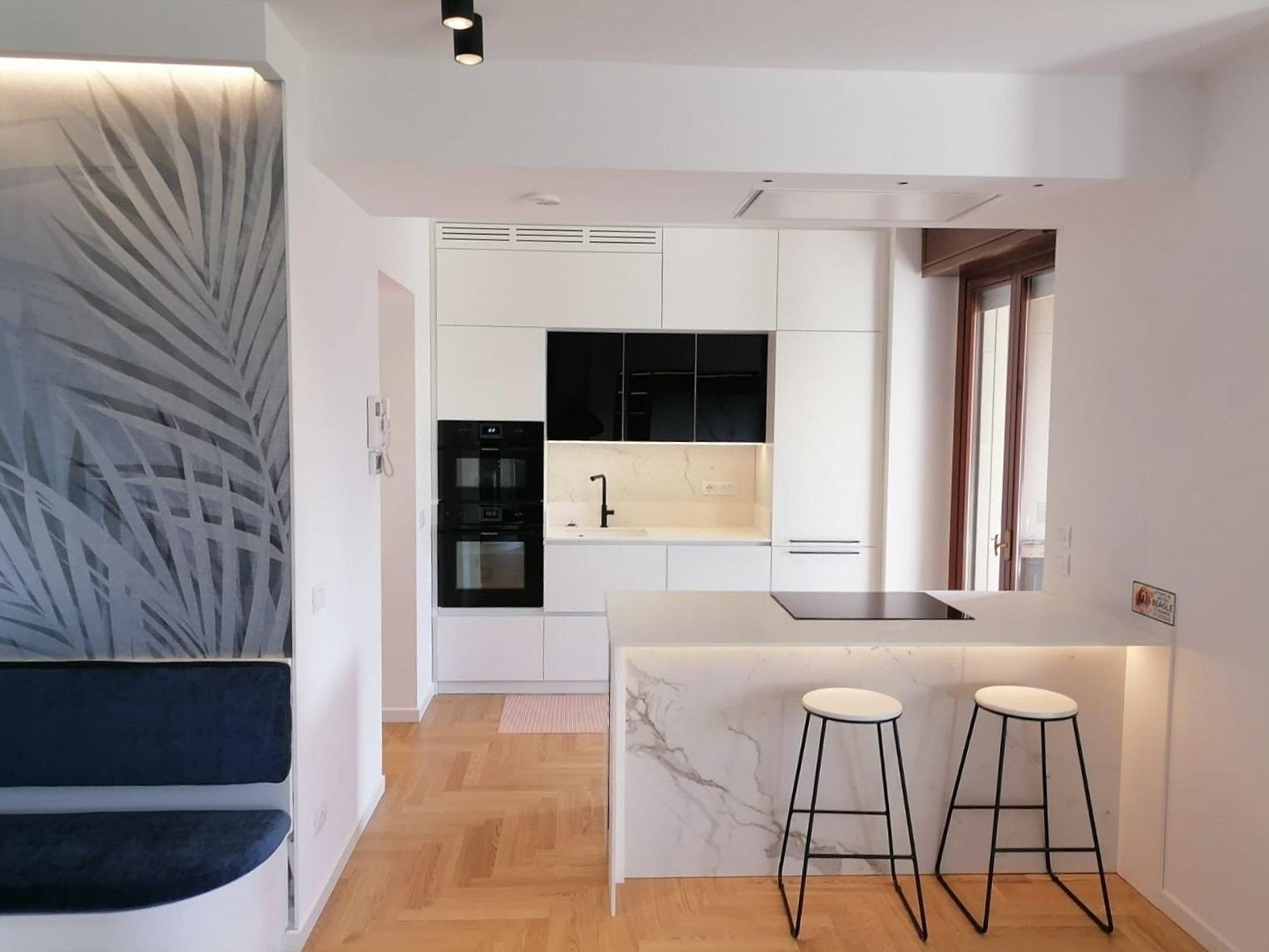 Cucina moderna bianca con isola in marmo, due sgabelli, panca blu e decorazione murale con motivo a foglie.