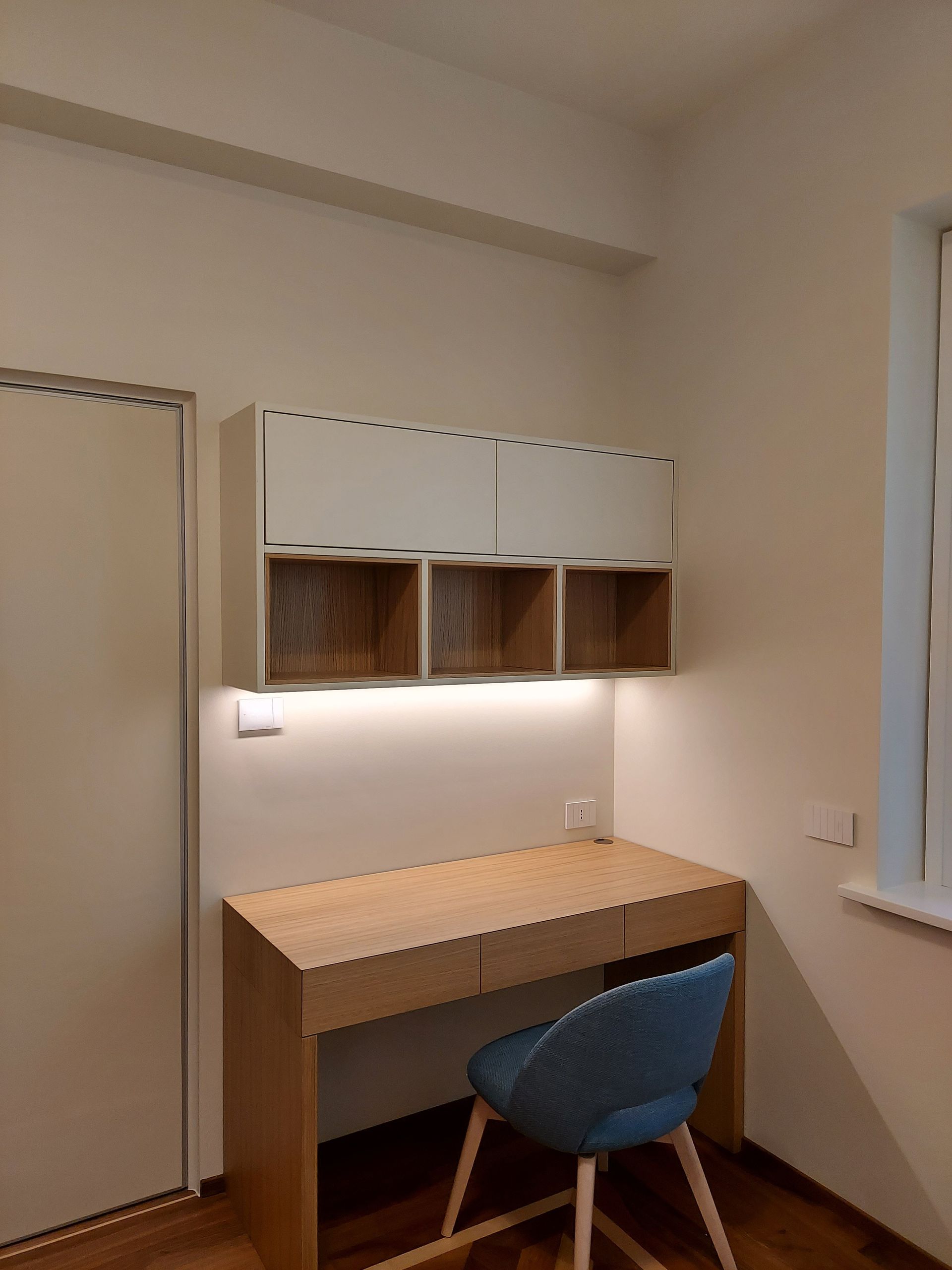 Piccolo angolo studio con scrivania in legno, sedia blu, armadietto a muro e luce soffusa sotto i pensili.
