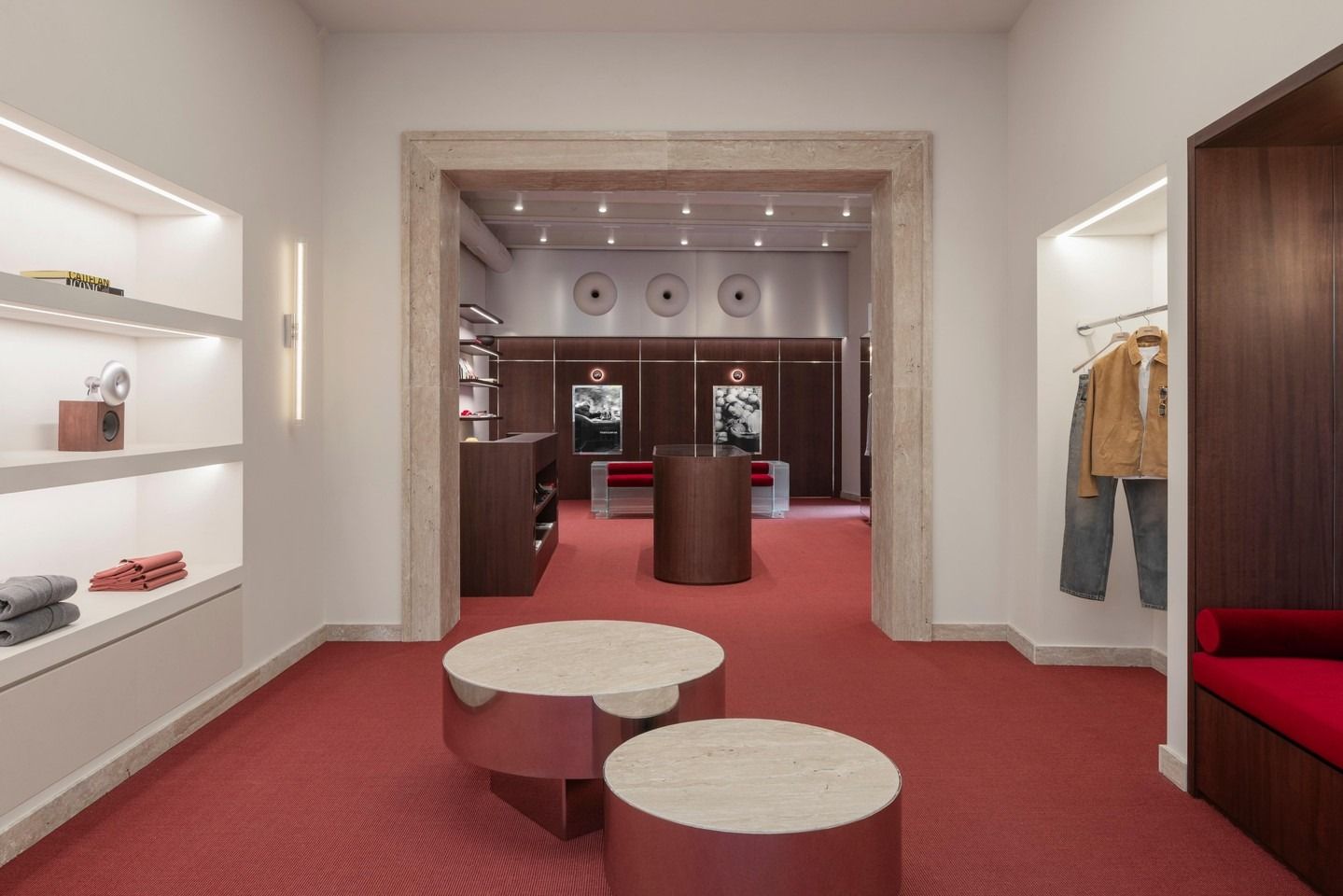 Boutique di abbigliamento moderna con tappeto rosso, scaffali espositivi bianchi e tavolini rotondi al centro.