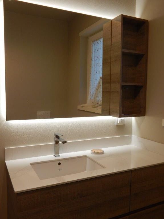 Mobile bagno moderno con lavabo bianco, ante in legno, specchio e illuminazione sottopensile dai toni caldi.