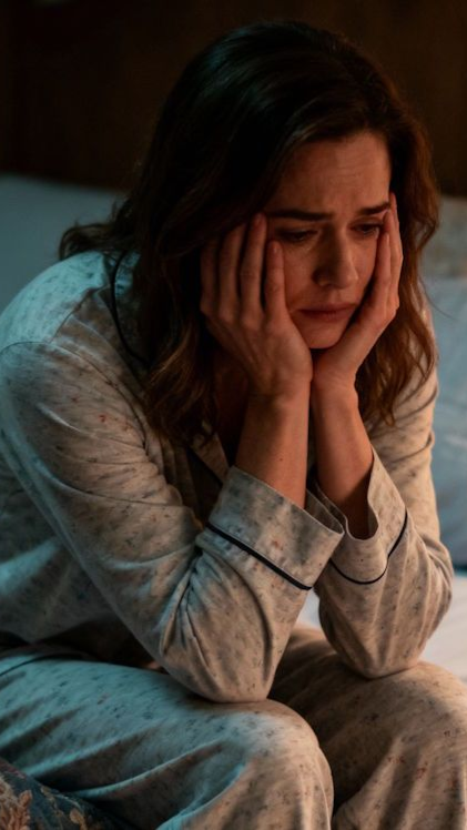 A tired OKC woman in her pj's sitting on the edge of her bed hands covering face with a warm beside lamp, pillow and blanket