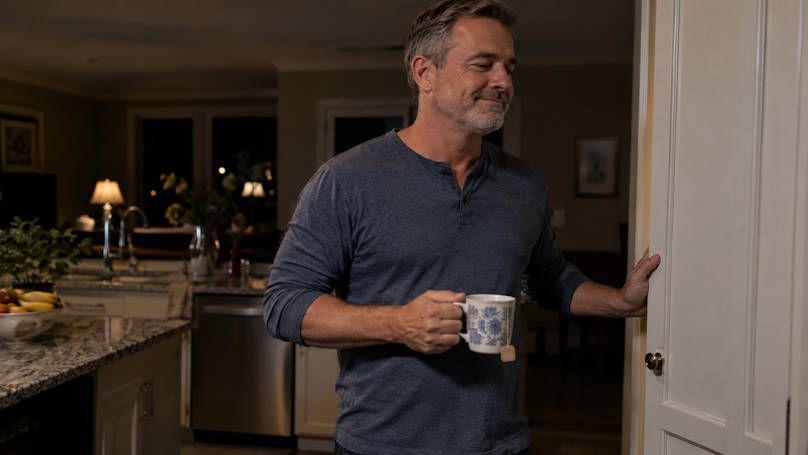 Man waking up sipping a cup of tea with snack cupboard open with a smile on his face