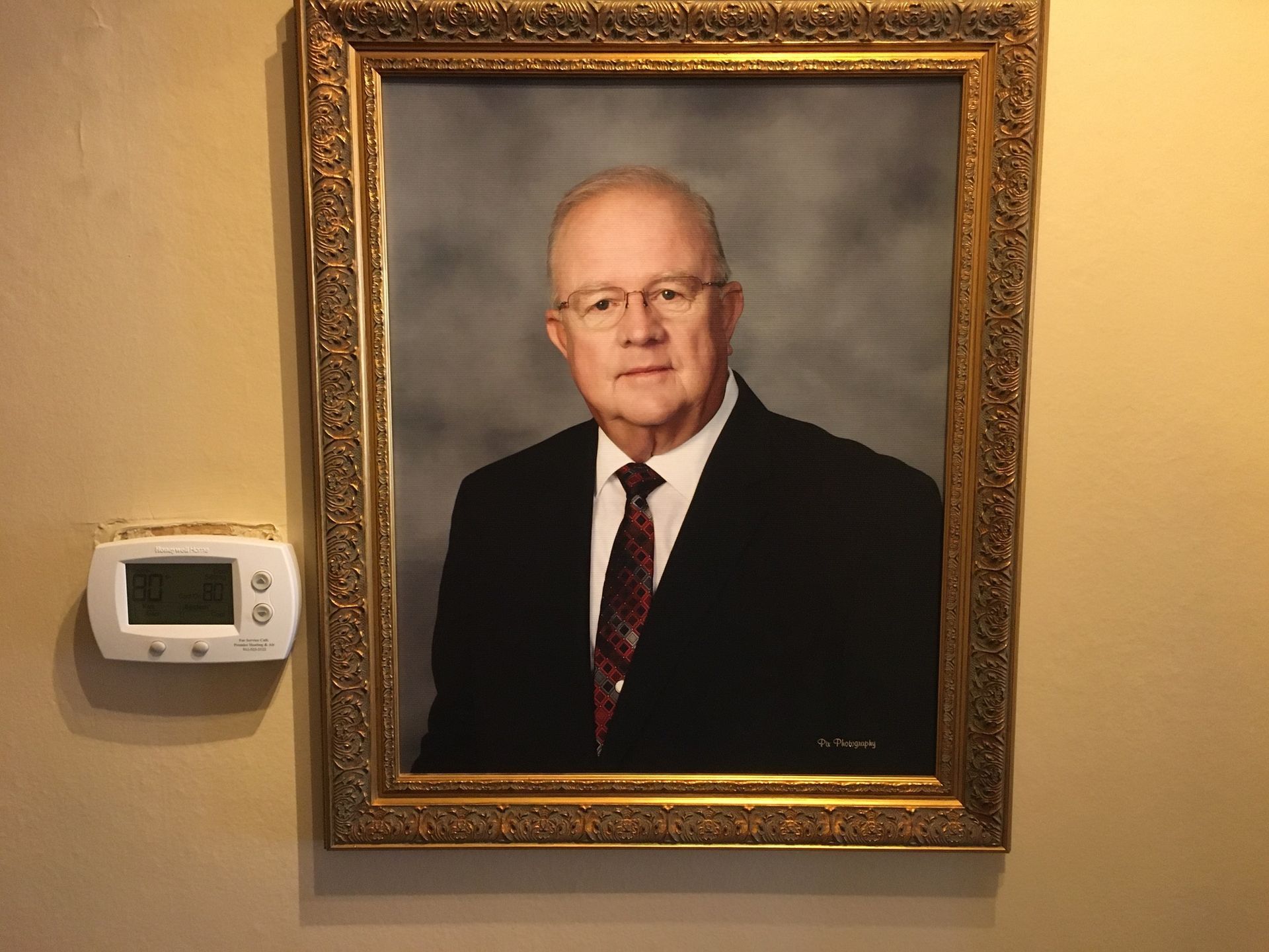 Framed portrait of a man in a suit hanging on a beige wall beside a thermostat.