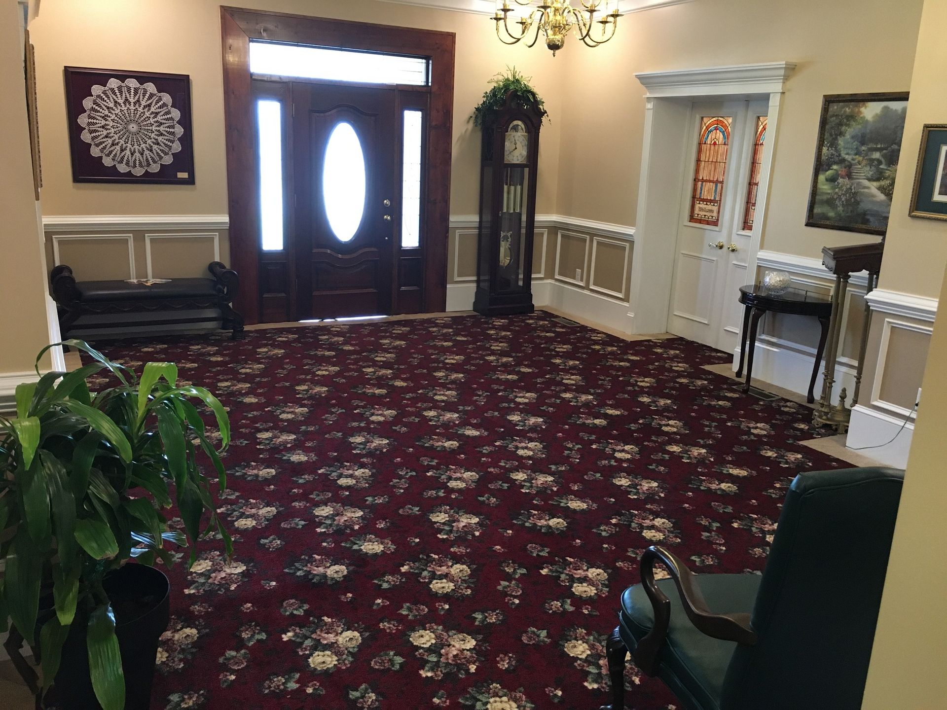 Carpeted lobby with double front doors, plants, chairs, and wall art under warm lighting