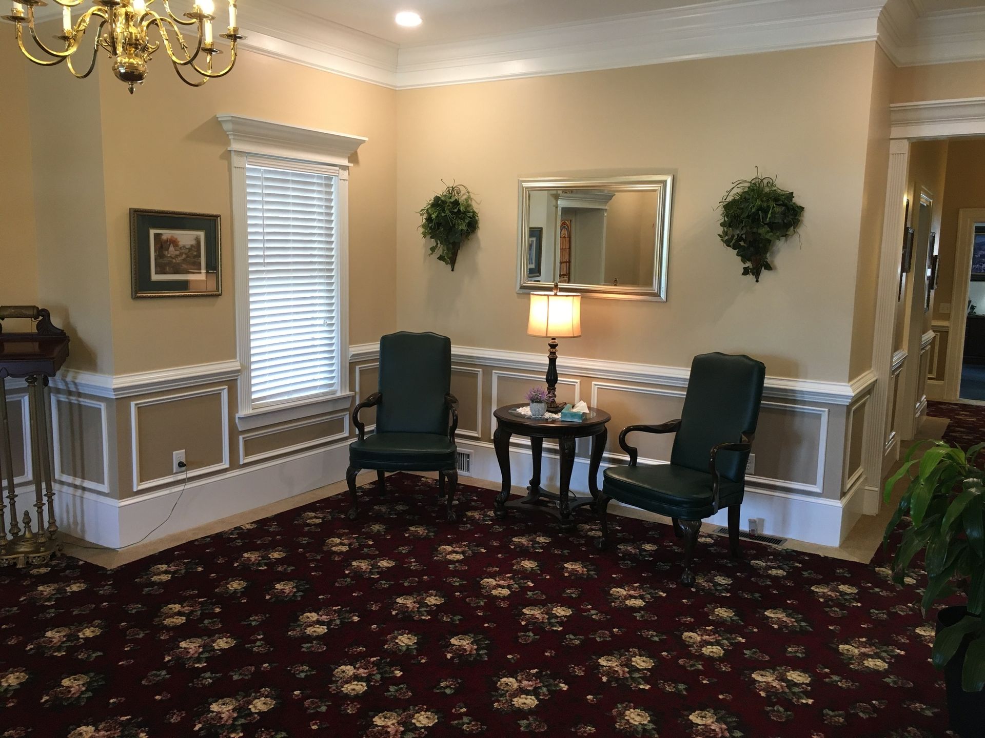 Small waiting room with two green chairs, round table, lamp, and floral carpet in beige walls.