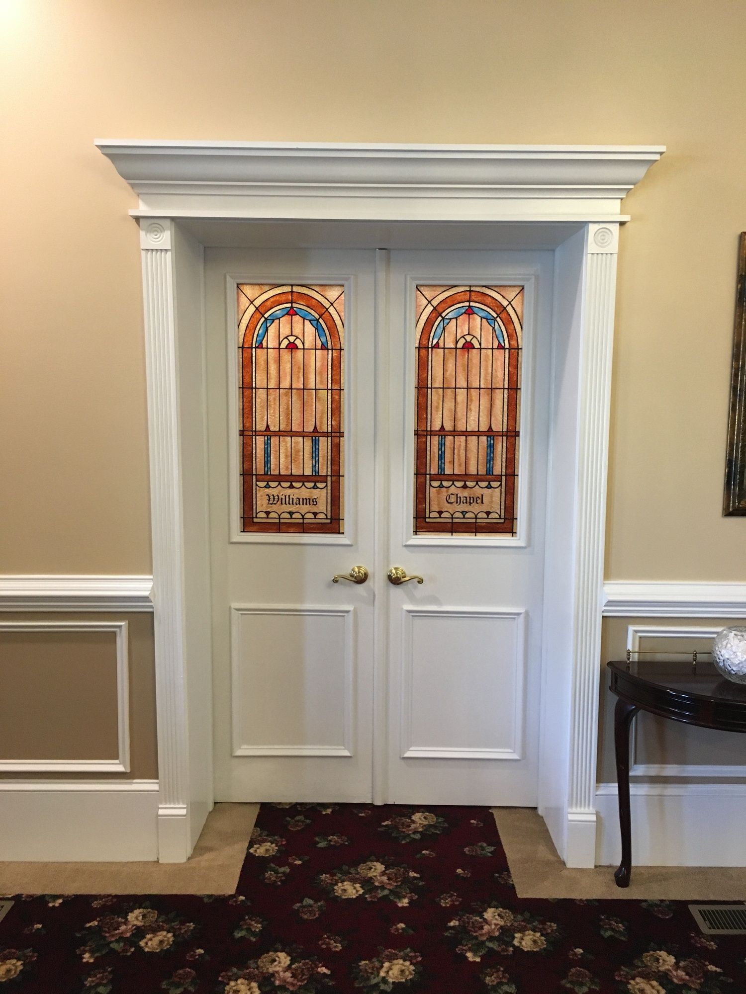 White double doors with stained-glass panels in a beige hallway with crown molding and a floral carpet.