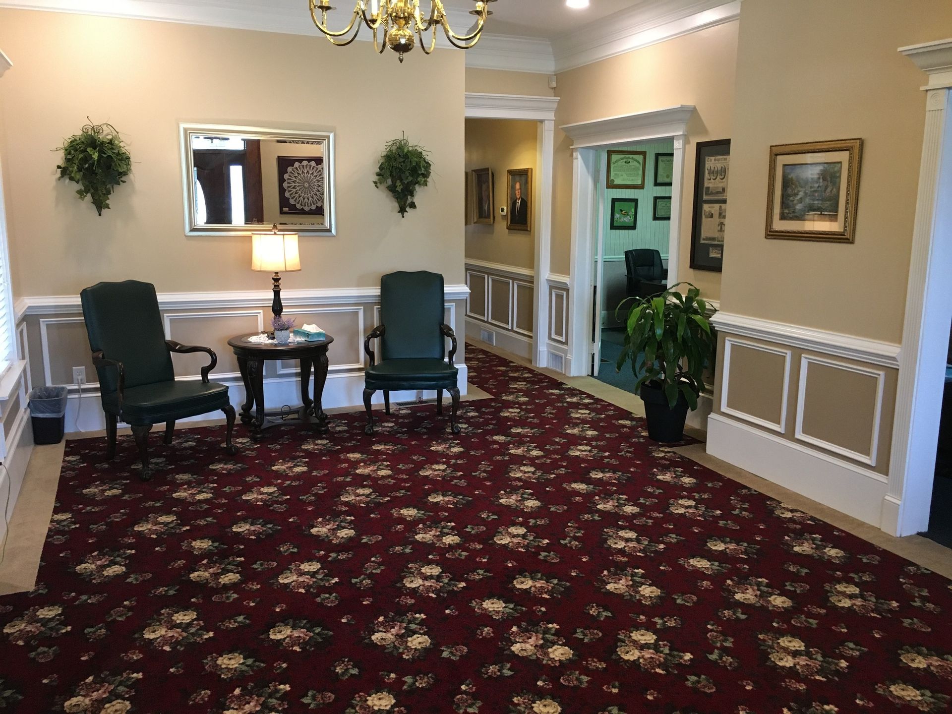 Empty hotel lobby with red patterned carpet, two green chairs, lamp, and framed wall art.