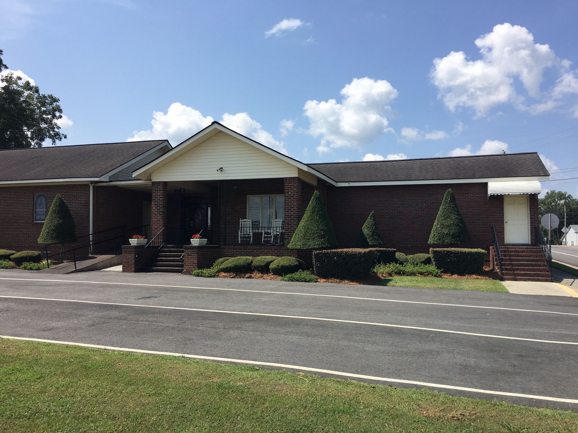 Single-story brick building with a white gabled entrance, shrubs, and a paved driveway under a blue sky.