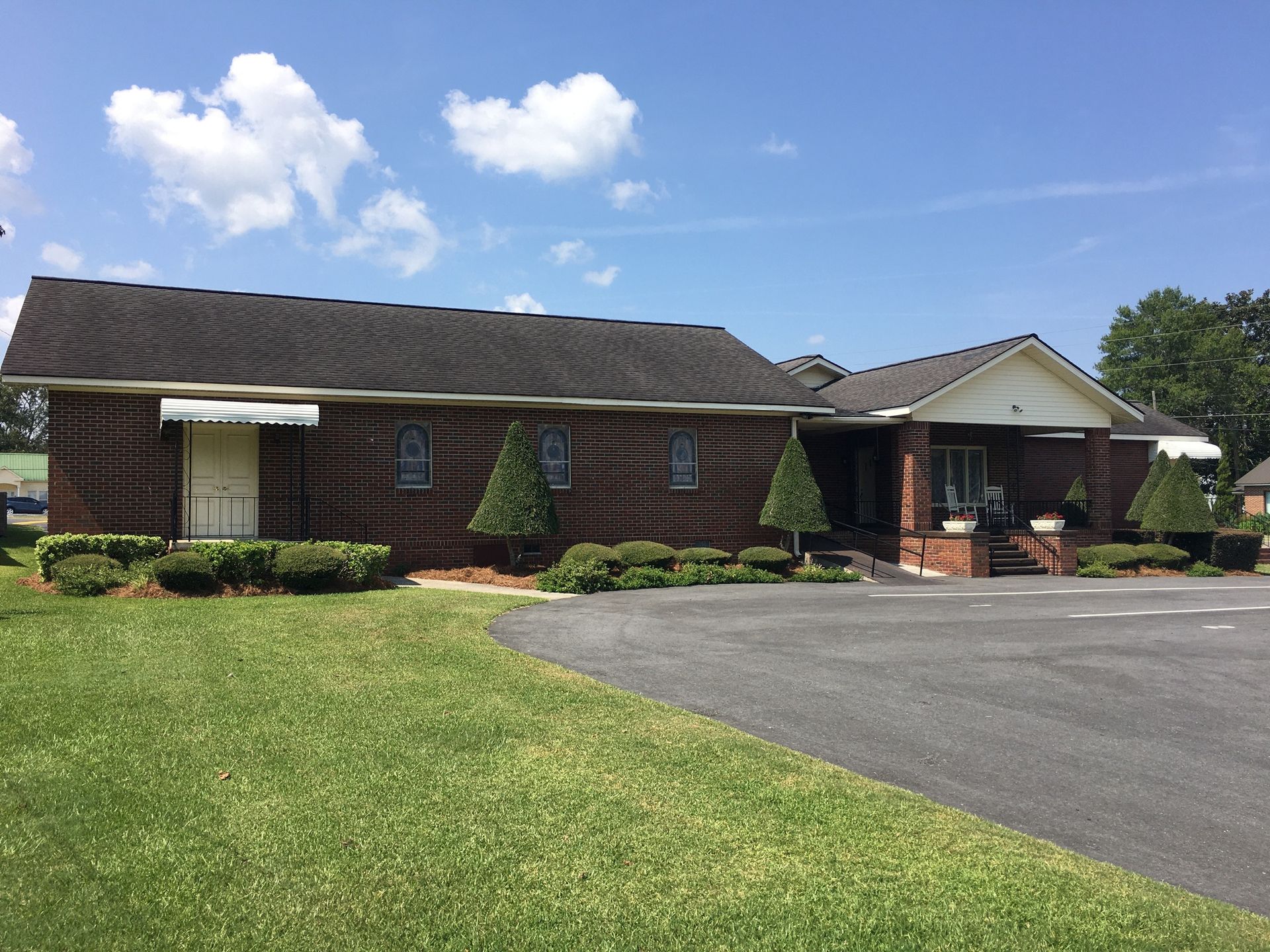Single-story brick house with a driveway, front porch, and a large green lawn under a blue sky.