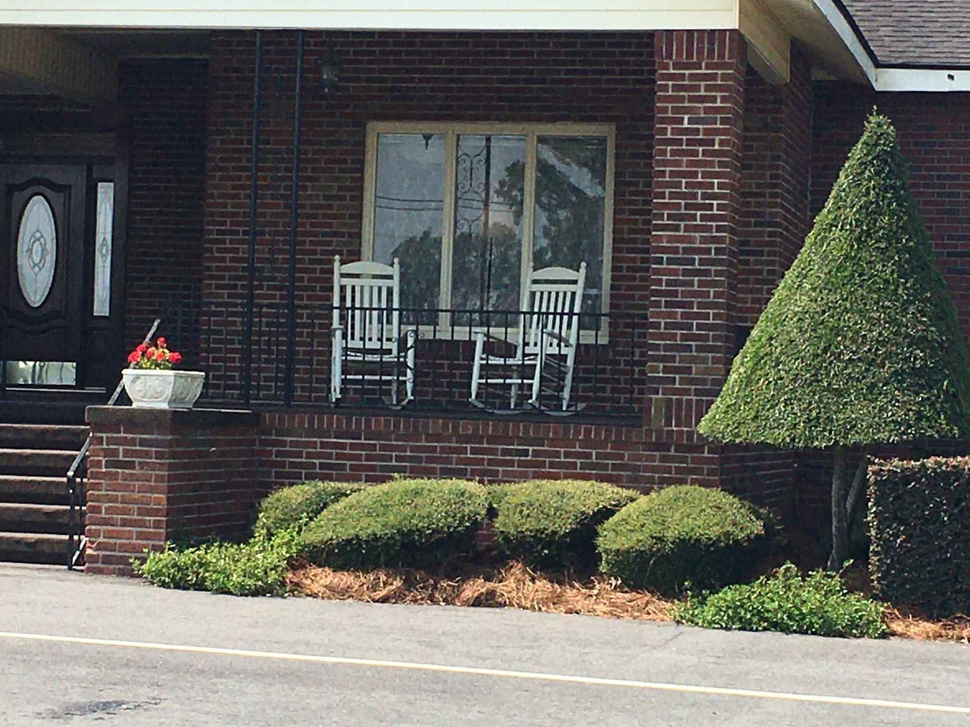 Brick house front porch with white chairs, potted flowers, and trimmed shrubs by the driveway