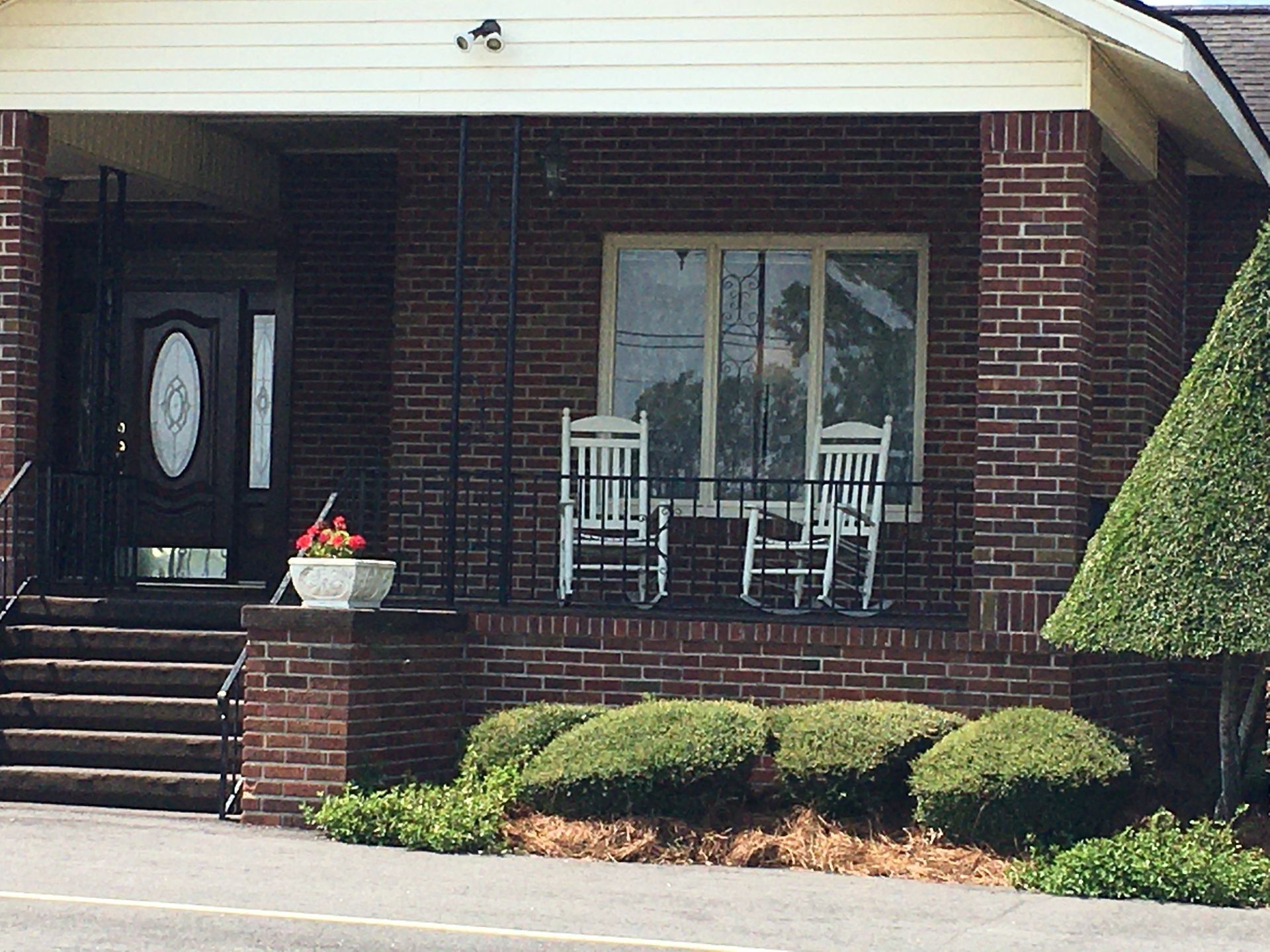 Brick house porch with white rocking chairs, front steps, shrubs, and a glass door