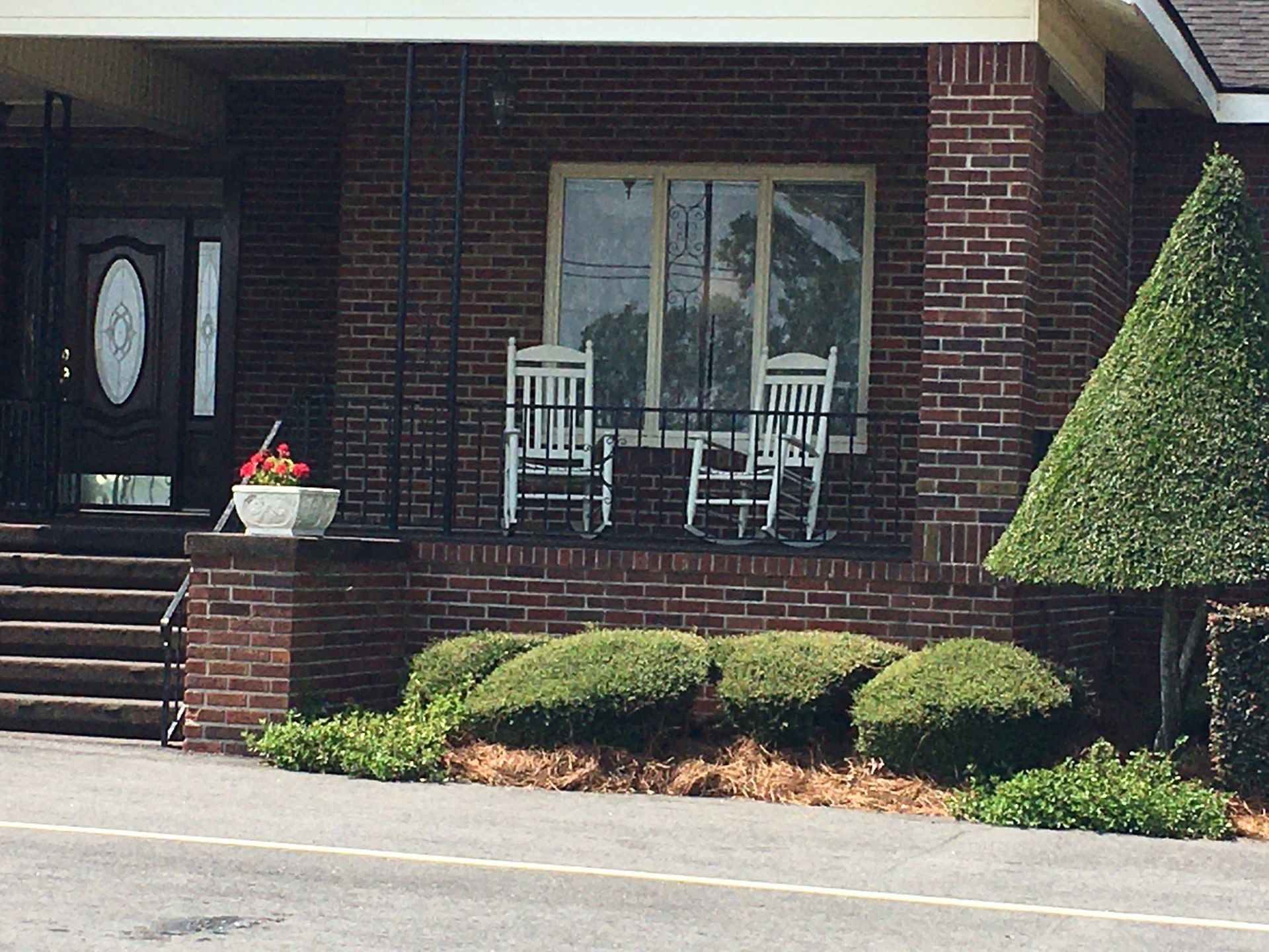 Front porch of a brick house with a white rocking chair, window, steps, and trimmed shrubs