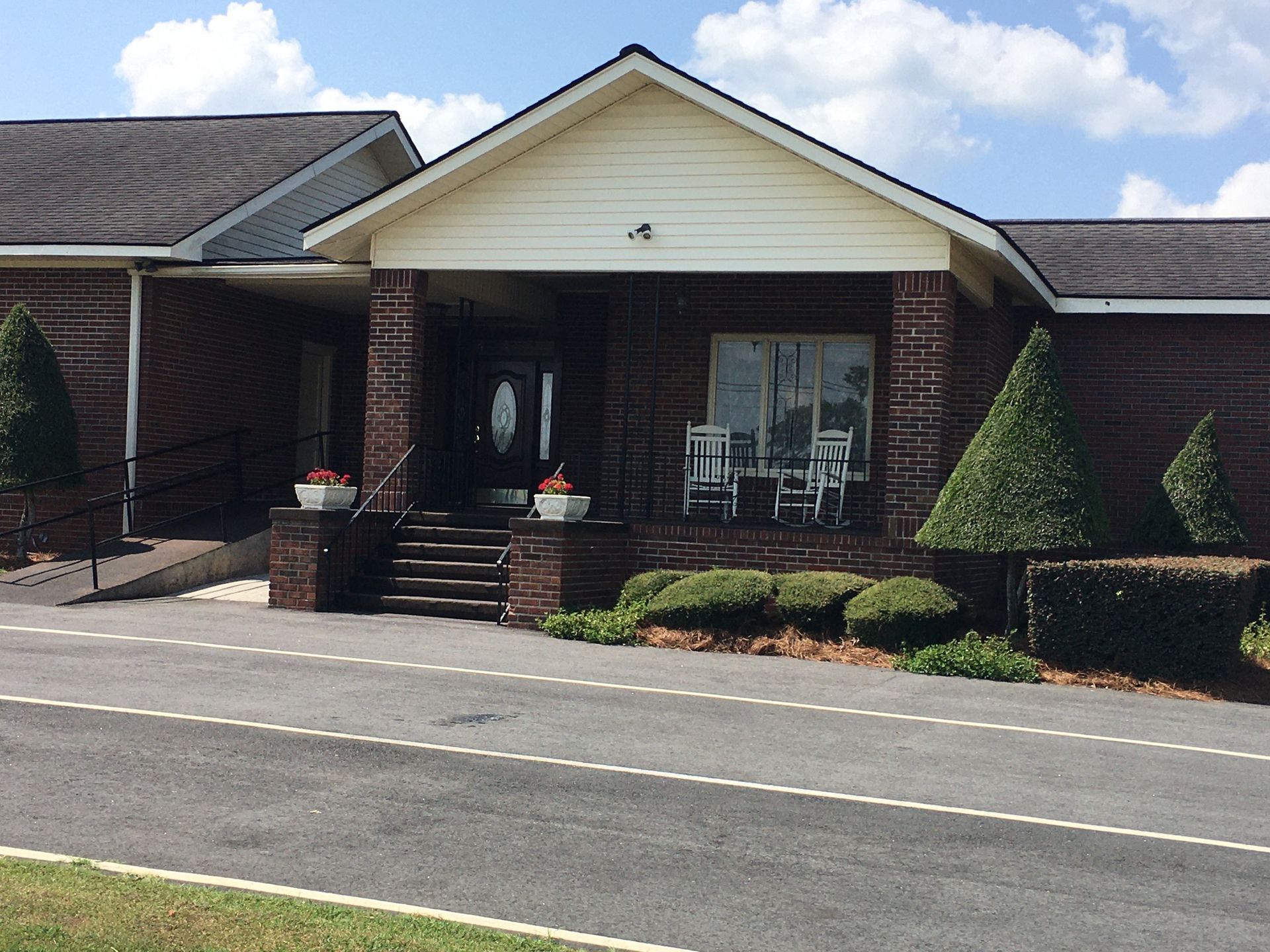 Front entrance of a brick building with white gable siding, steps, shrubs, and a parking lot in front
