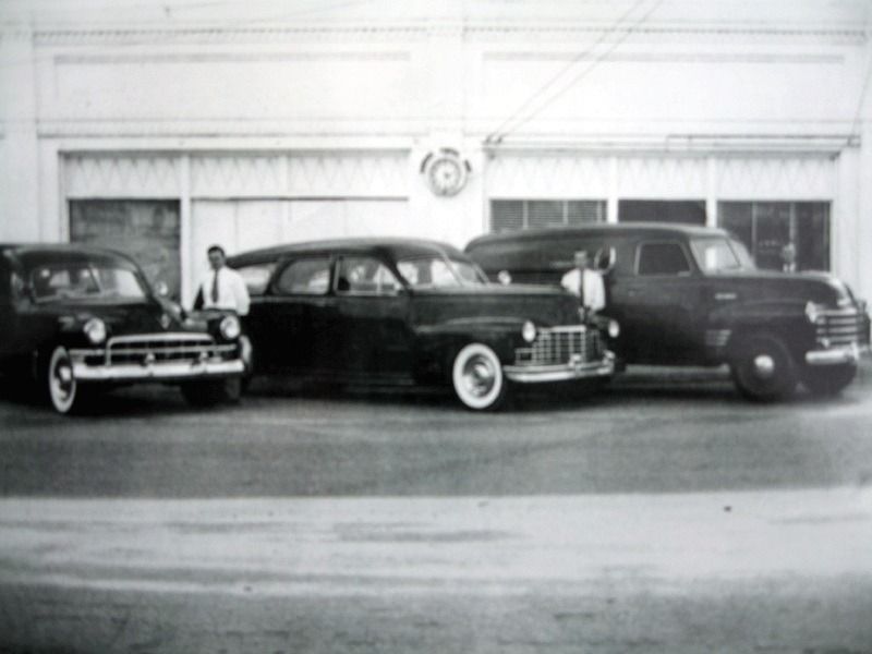 Three vintage cars parked in front of a white building, one person standing by the left car.